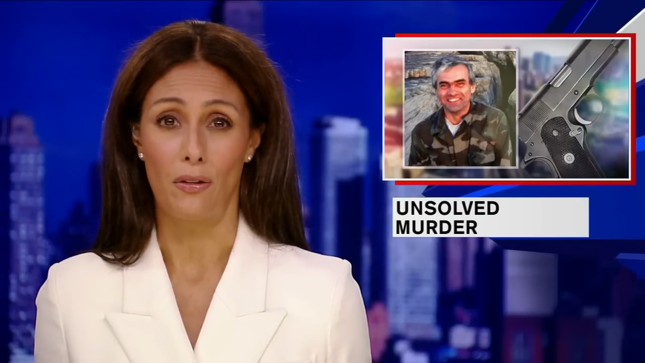 News anchor in white suit reports on an unsolved murder with an image of a person in military-style clothing and a gun