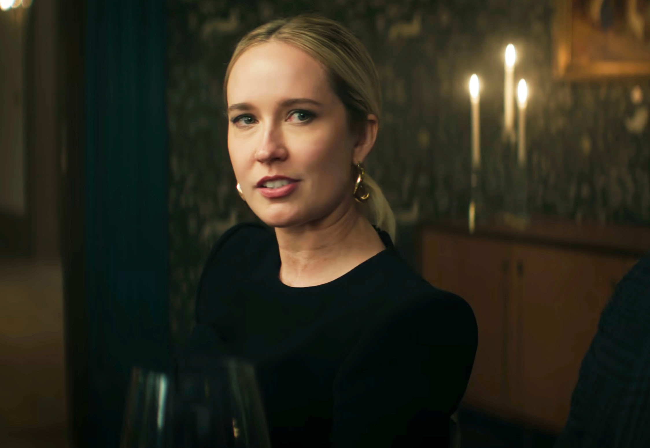 Person seated indoors, wearing elegant attire and earrings, with softly lit candles in the background