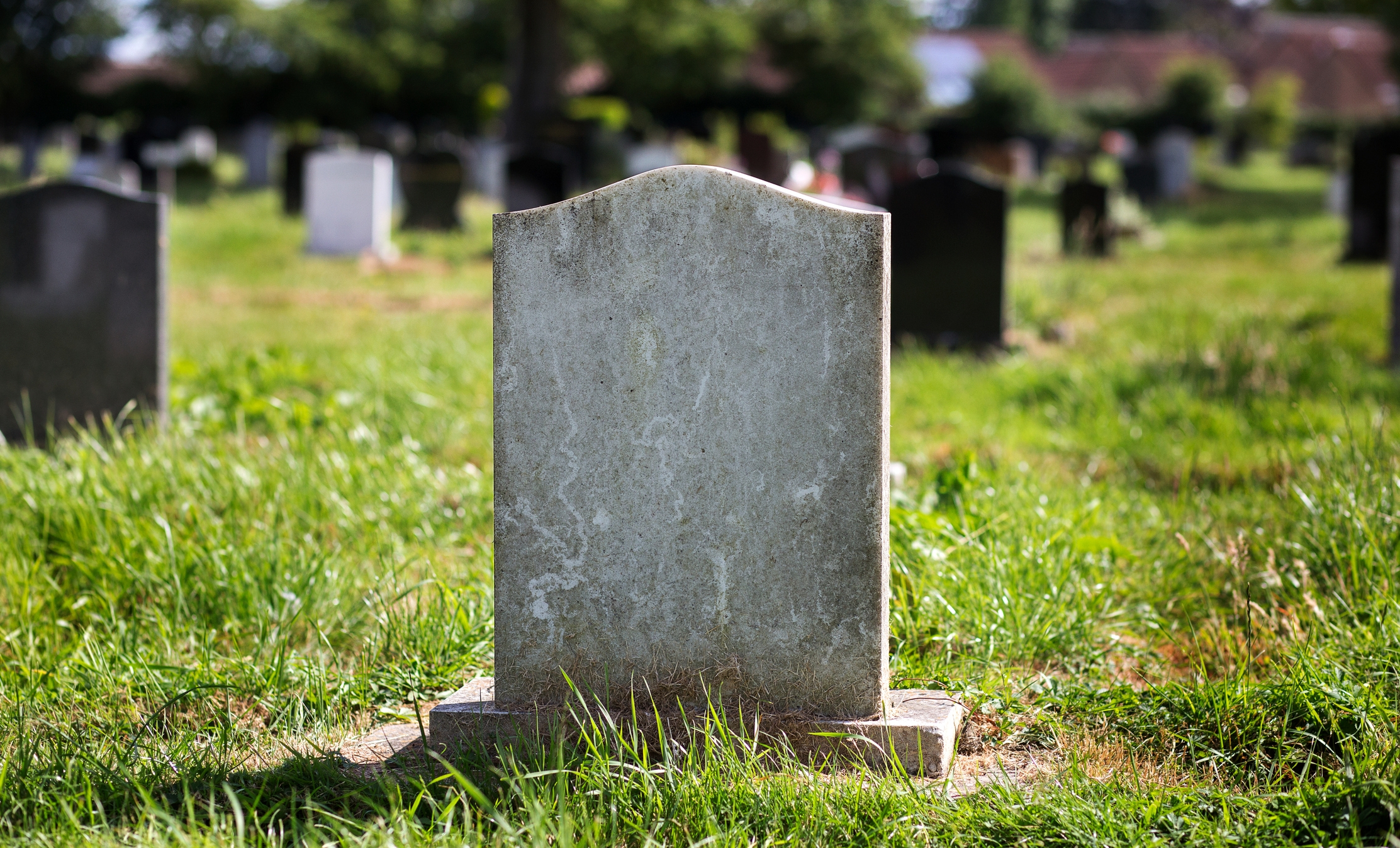 A weathered, blank tombstone stands in a grassy cemetery, surrounded by other headstones, with trees and a building in the background