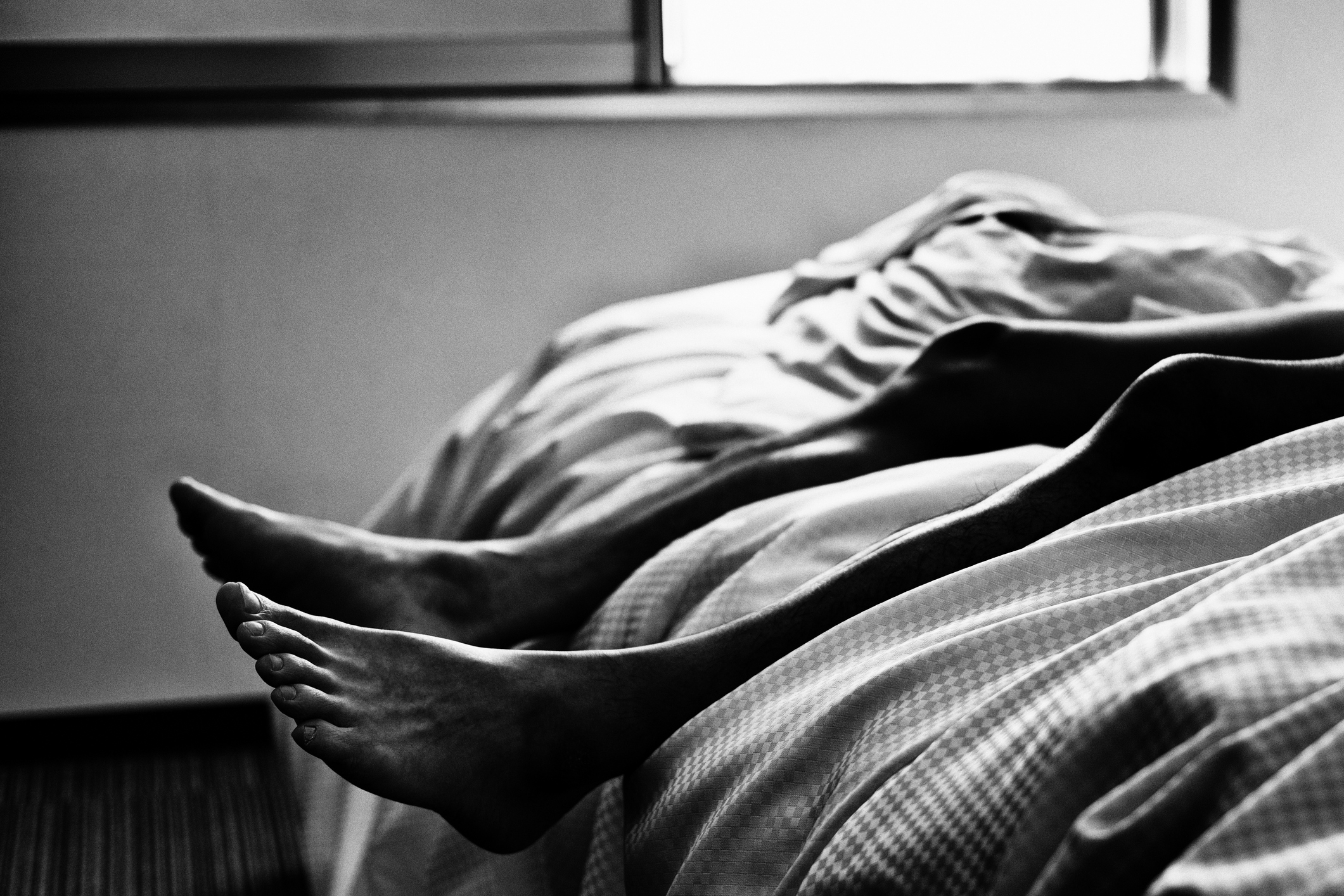 Person lying on bed with feet hanging off the edge, under a crumpled sheet, in a dimly lit room