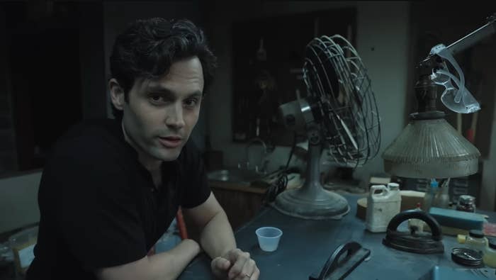 Person in a casual setting next to vintage fan and lamp, looking at the camera. Scene suggests a candid, informal moment