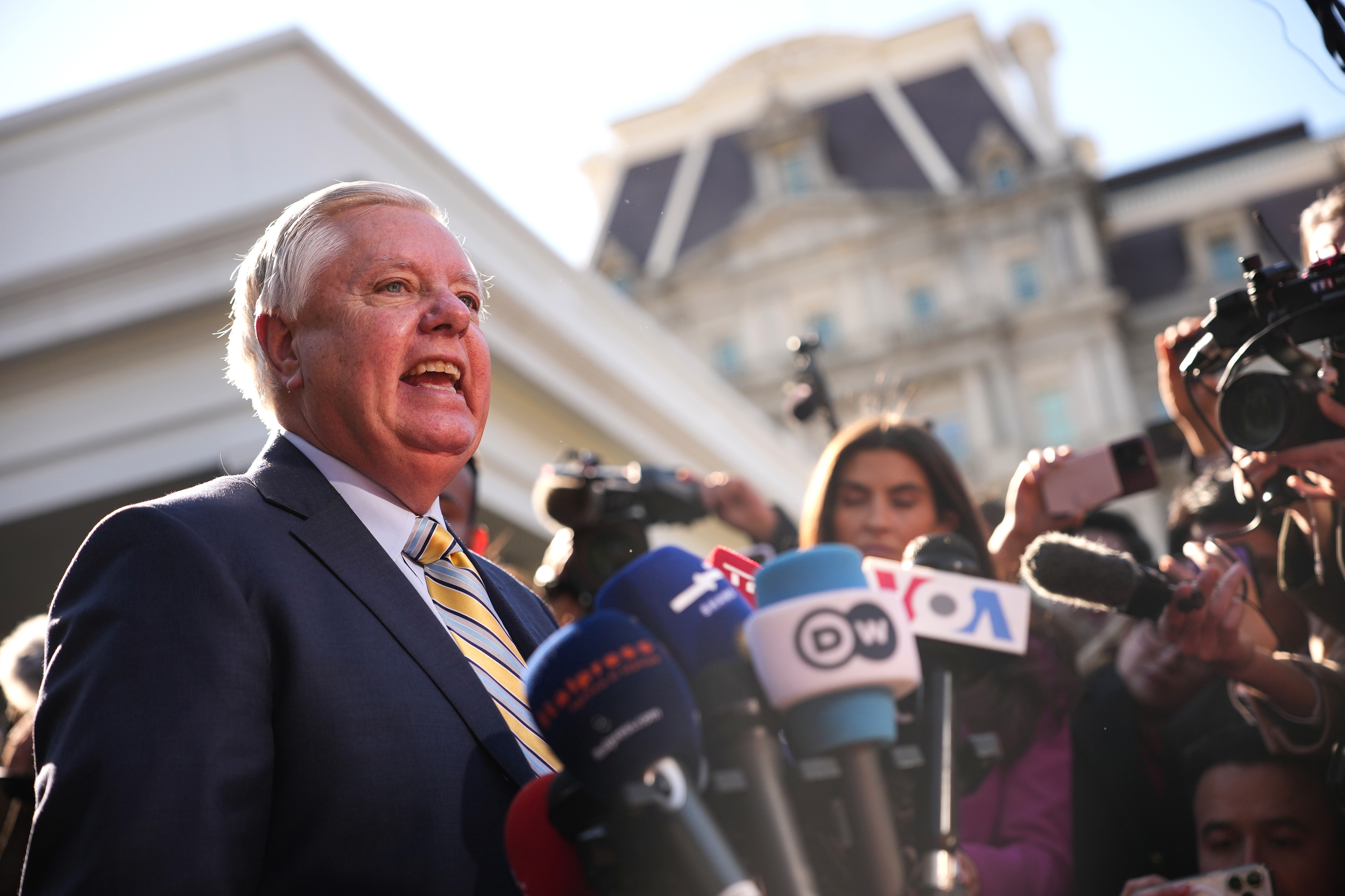 Person speaking at outdoor press conference, surrounded by microphones and reporters. Background shows a large building