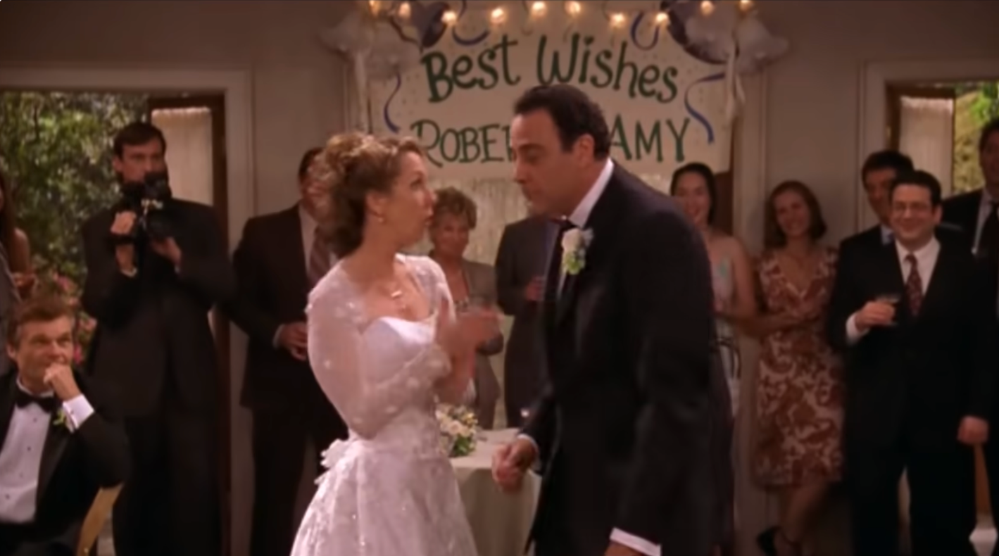 A bride in a lace wedding dress and a groom in a suit stand in front of a "Best Wishes Robert & Amy" sign at a wedding reception with guests applauding