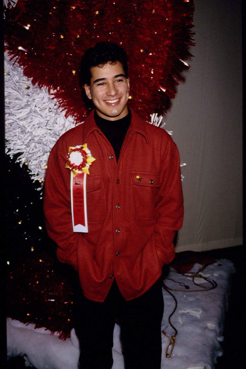 Person in a casual red jacket with an award ribbon, smiling against a festive backdrop of red and white decorations