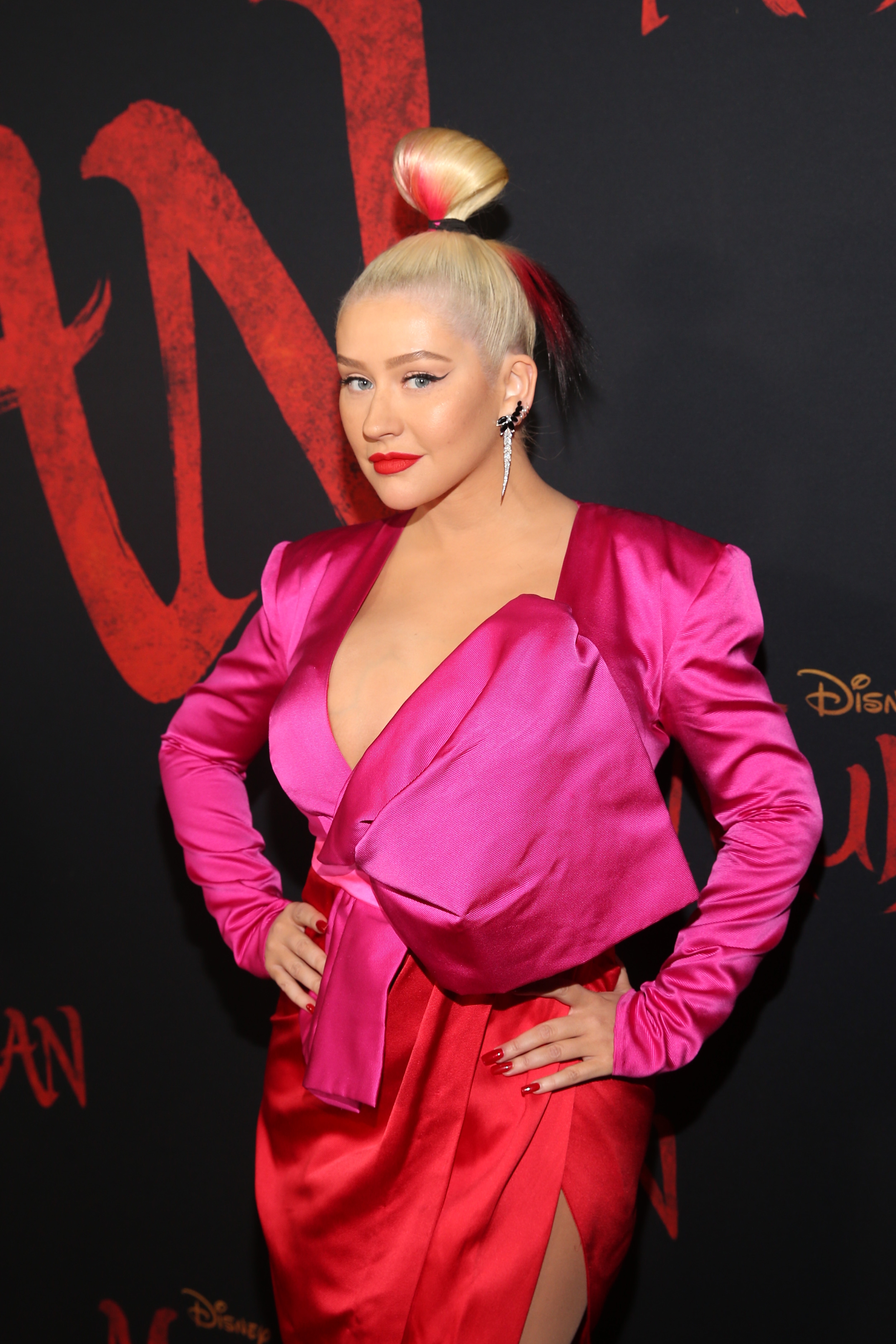 A person poses on the red carpet in a vibrant pink outfit with bold shoulder pads and a large bow, hair styled in a high bun