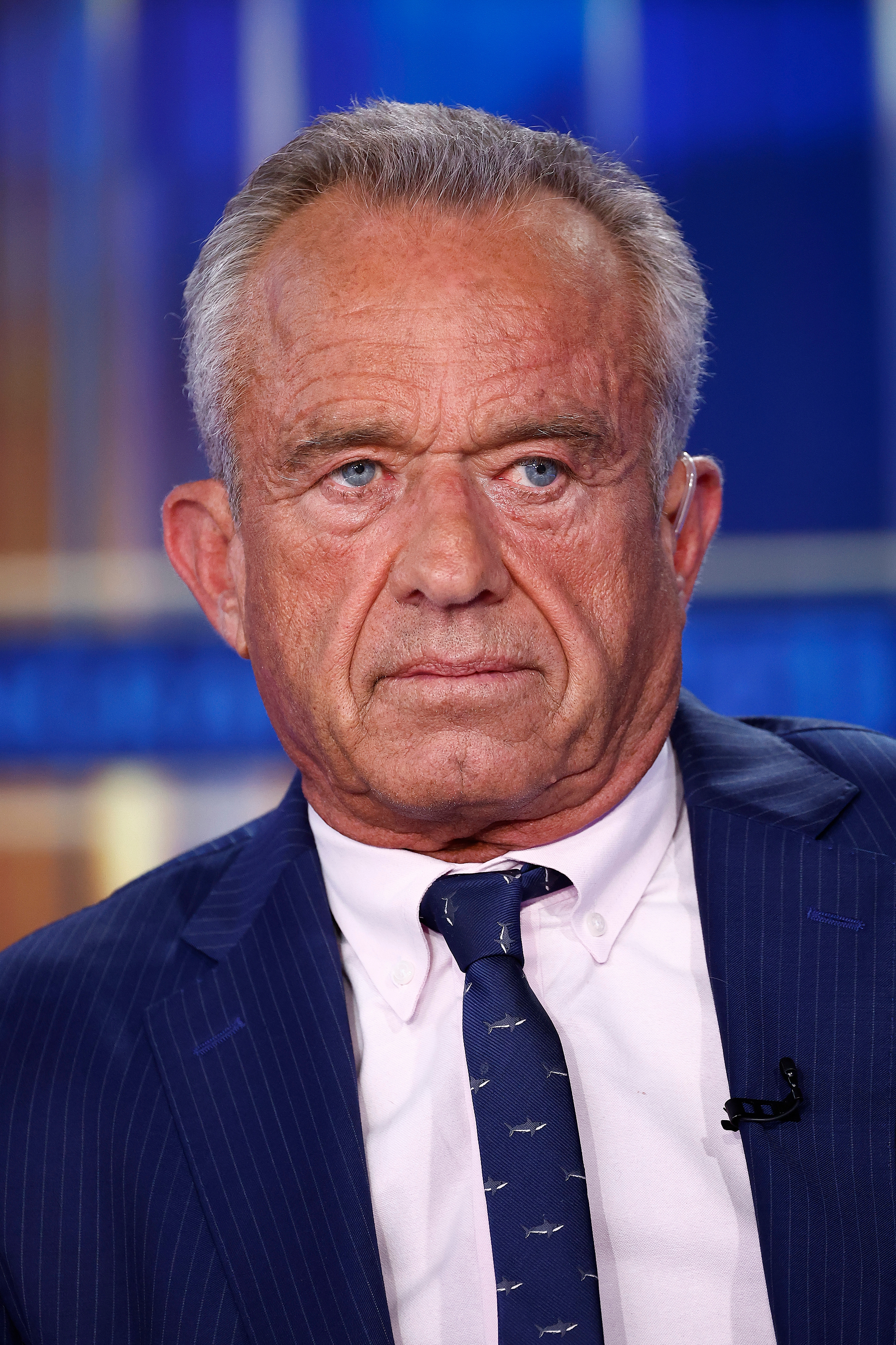 Older man in a suit and tie with a lapel microphone, appearing serious in a professional setting