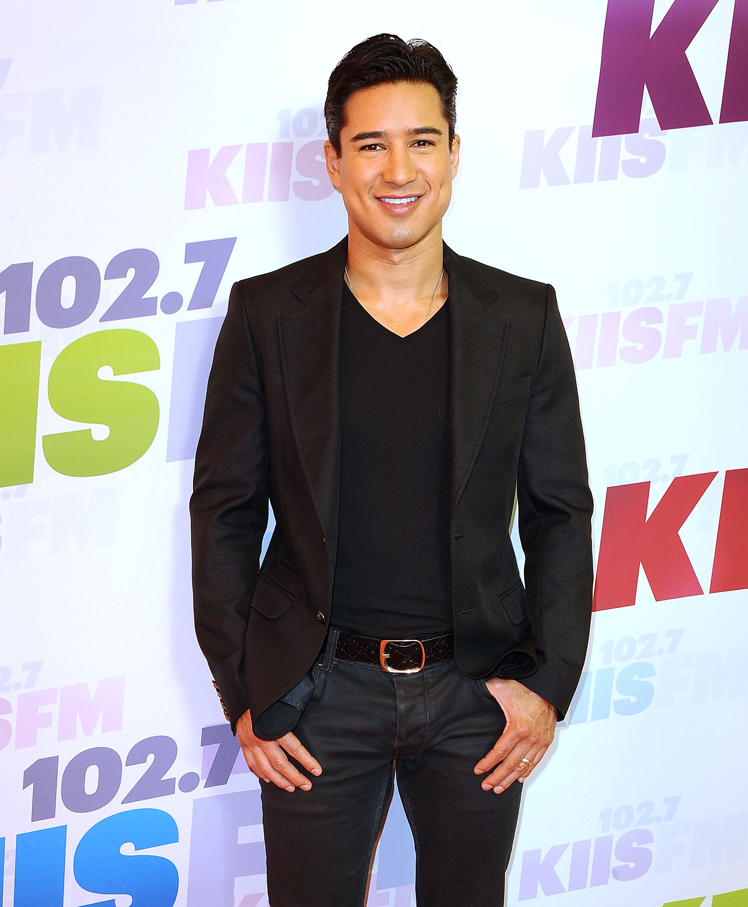 Celebrity posing on a red carpet in a fitted black blazer and v-neck shirt at a radio event backdrop