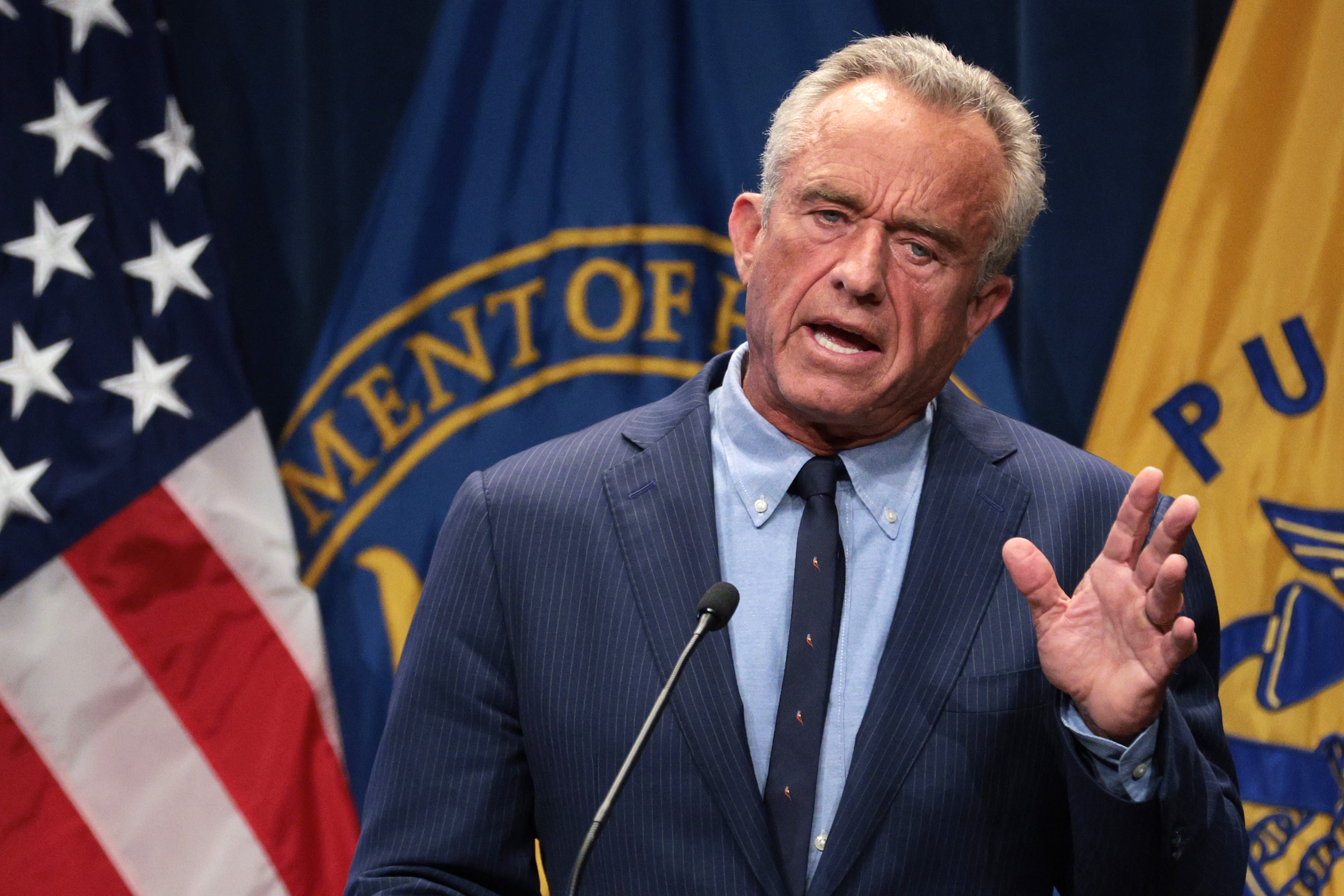 A man in a suit speaks at a podium with flags displaying government seals in the background
