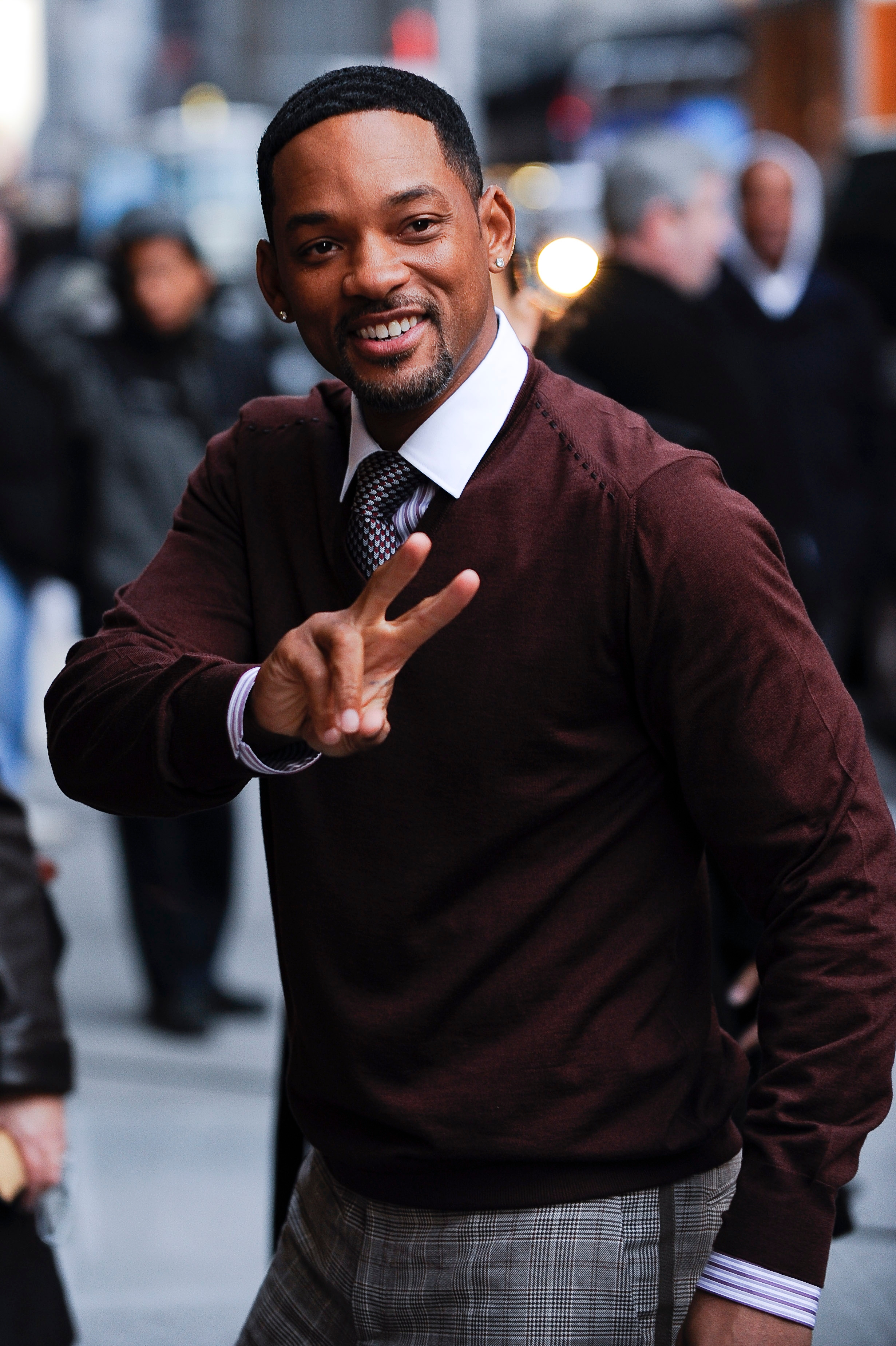 Person in a sweater and checkered pants flashes a peace sign on a busy street