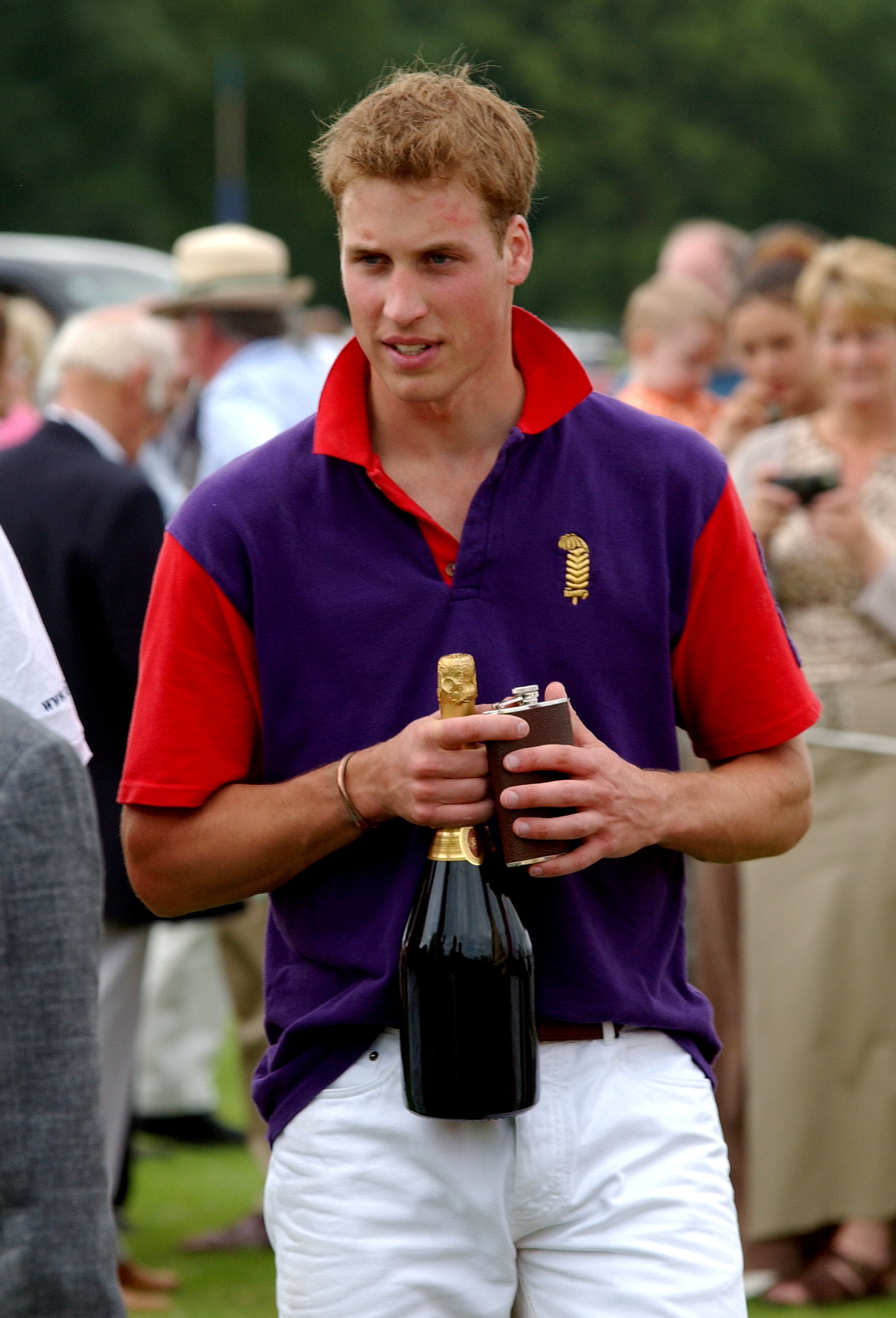 Person in a purple and red polo shirt and white pants holding a champagne bottle at an outdoor event