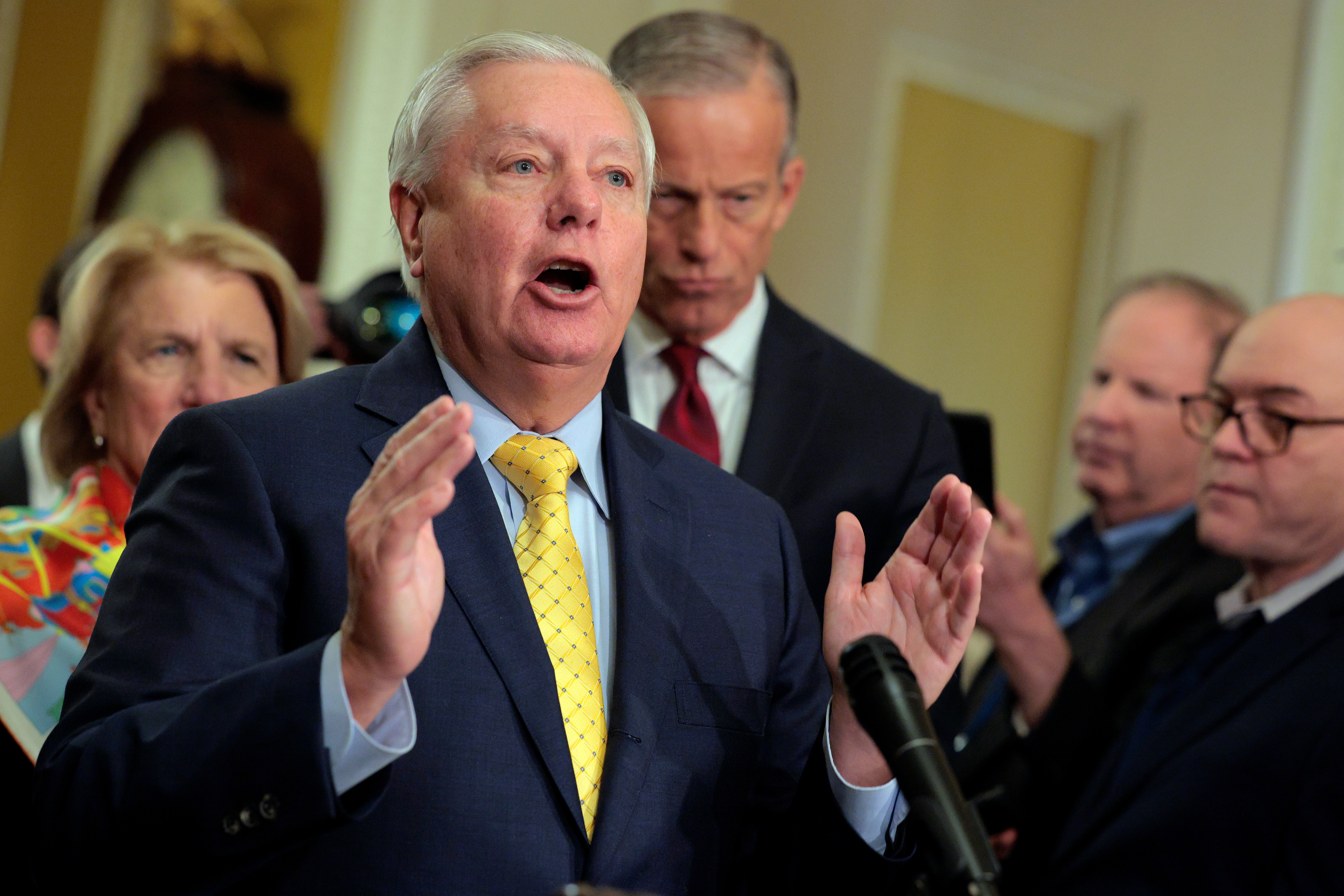 A person in a suit and tie speaks passionately during a media event, surrounded by several attentive individuals