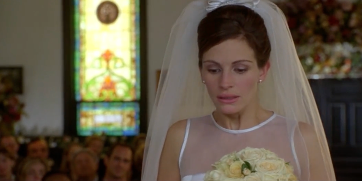 Bride in elegant wedding gown holding a bouquet, standing inside a church with a stained glass window in the background