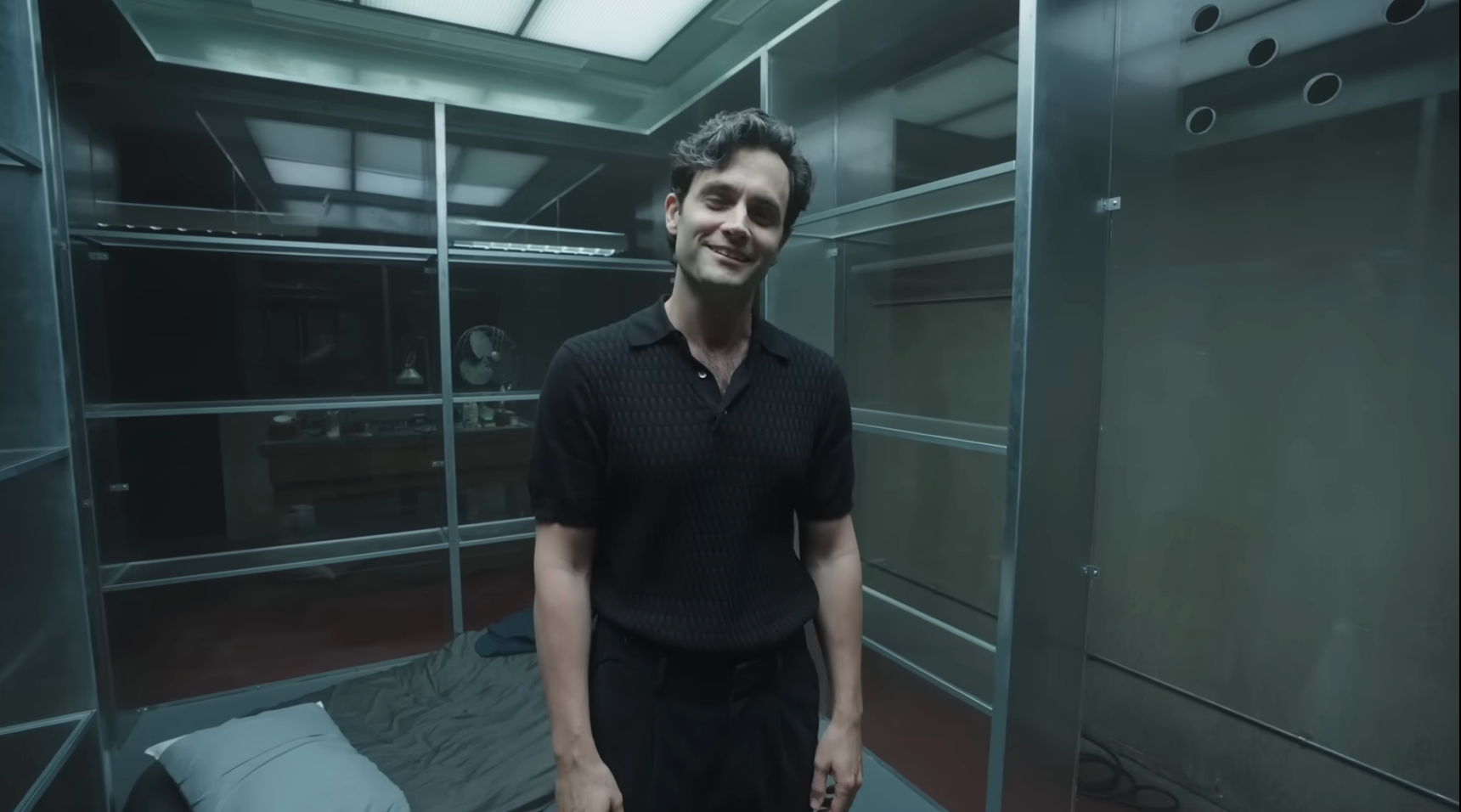 Person standing in minimalist room with glass walls and a bed, wearing a casual black outfit, smiling at the camera