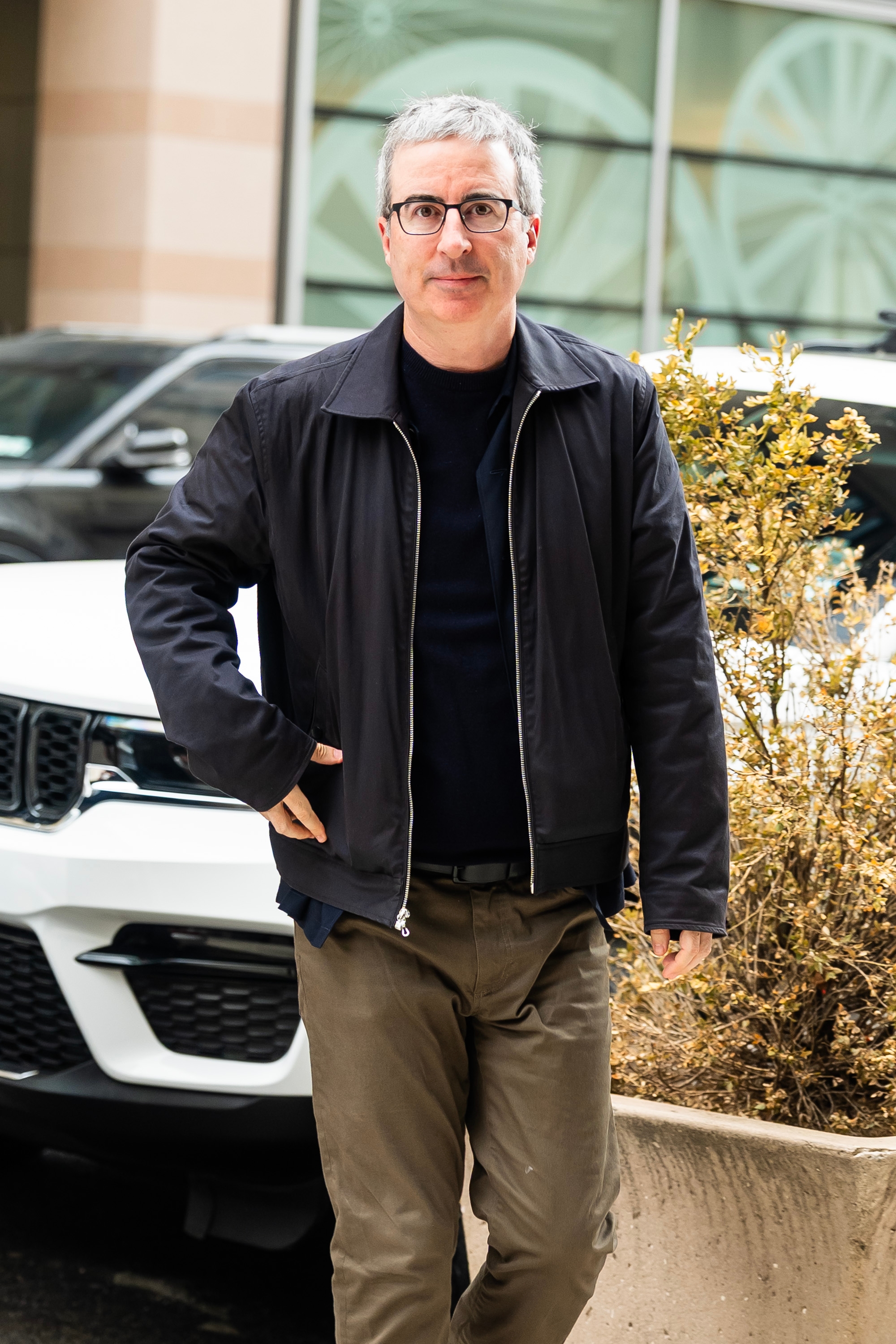 A person with short hair, glasses, a dark jacket, and khaki pants walks outdoors near a white car and a plant