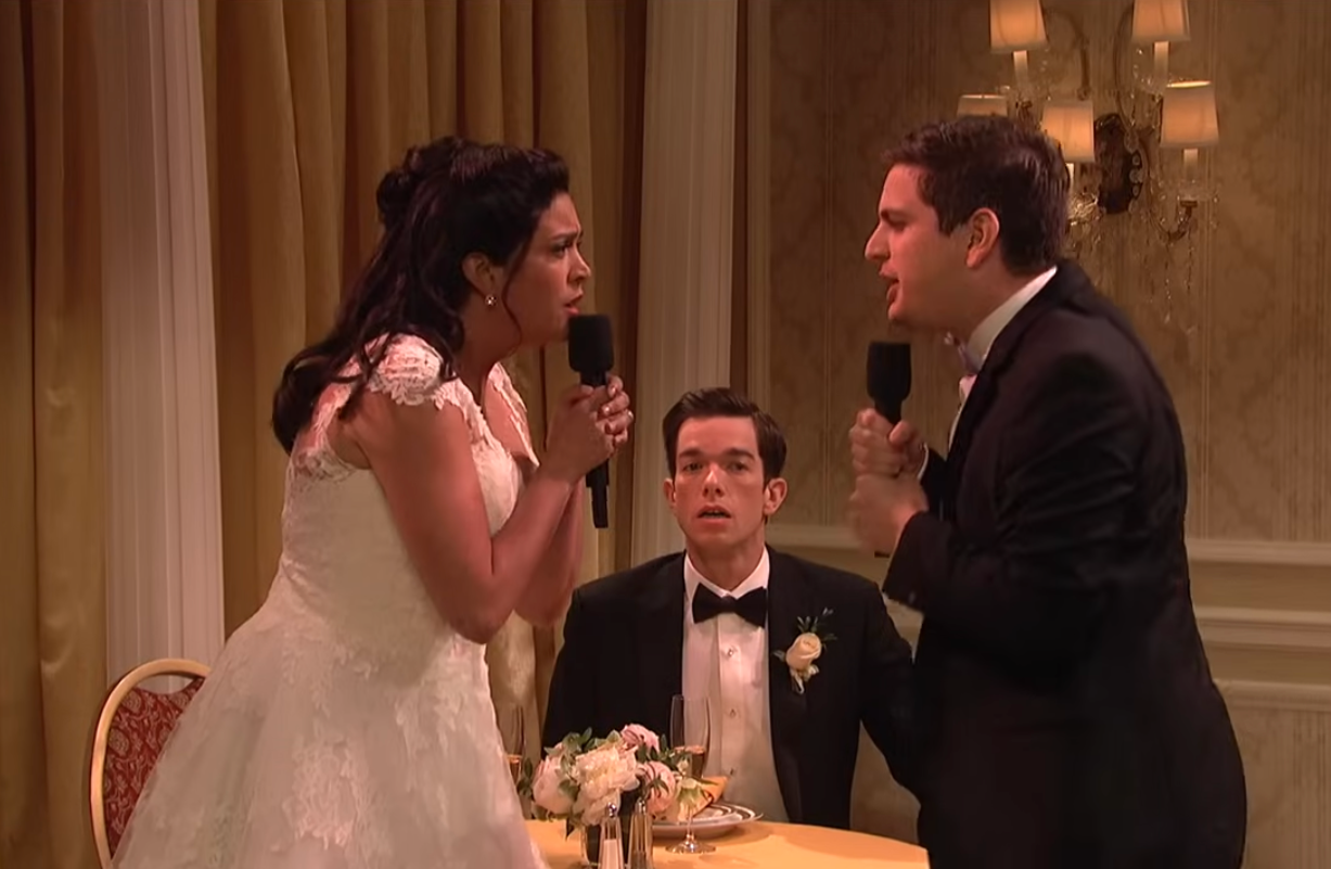 Bride and best man humorously sing into microphones at wedding reception; a groom in a tuxedo looks surprised