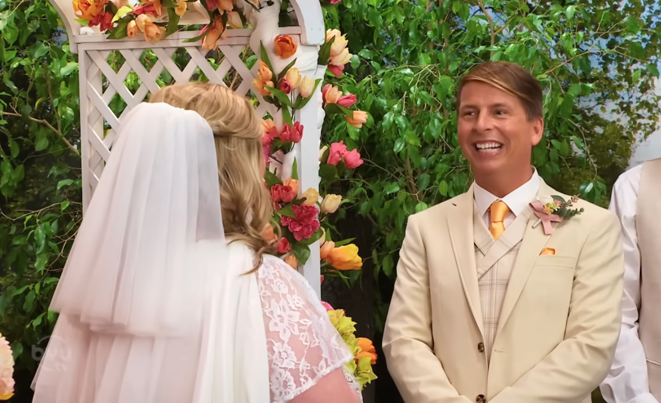 Wedding ceremony scene with a bride in lace and veil facing a groom in a suit with a tie under a floral archway