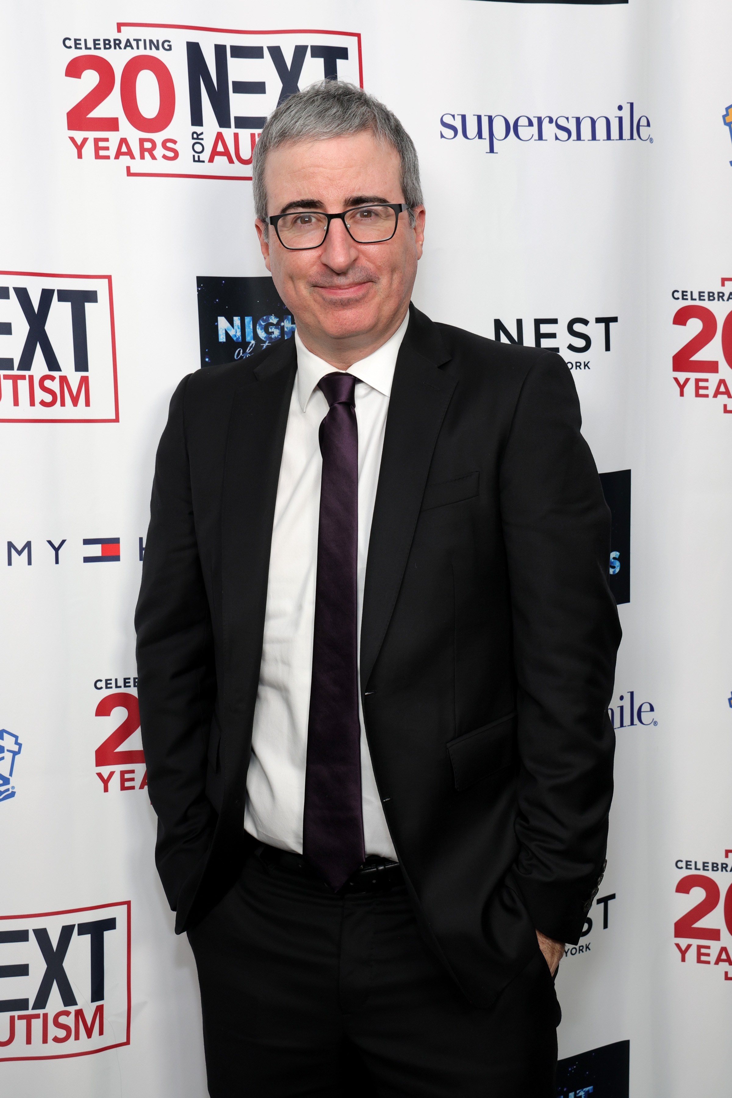 A person in a formal suit stands at a "Next for Autism" event backdrop, smiling at the camera