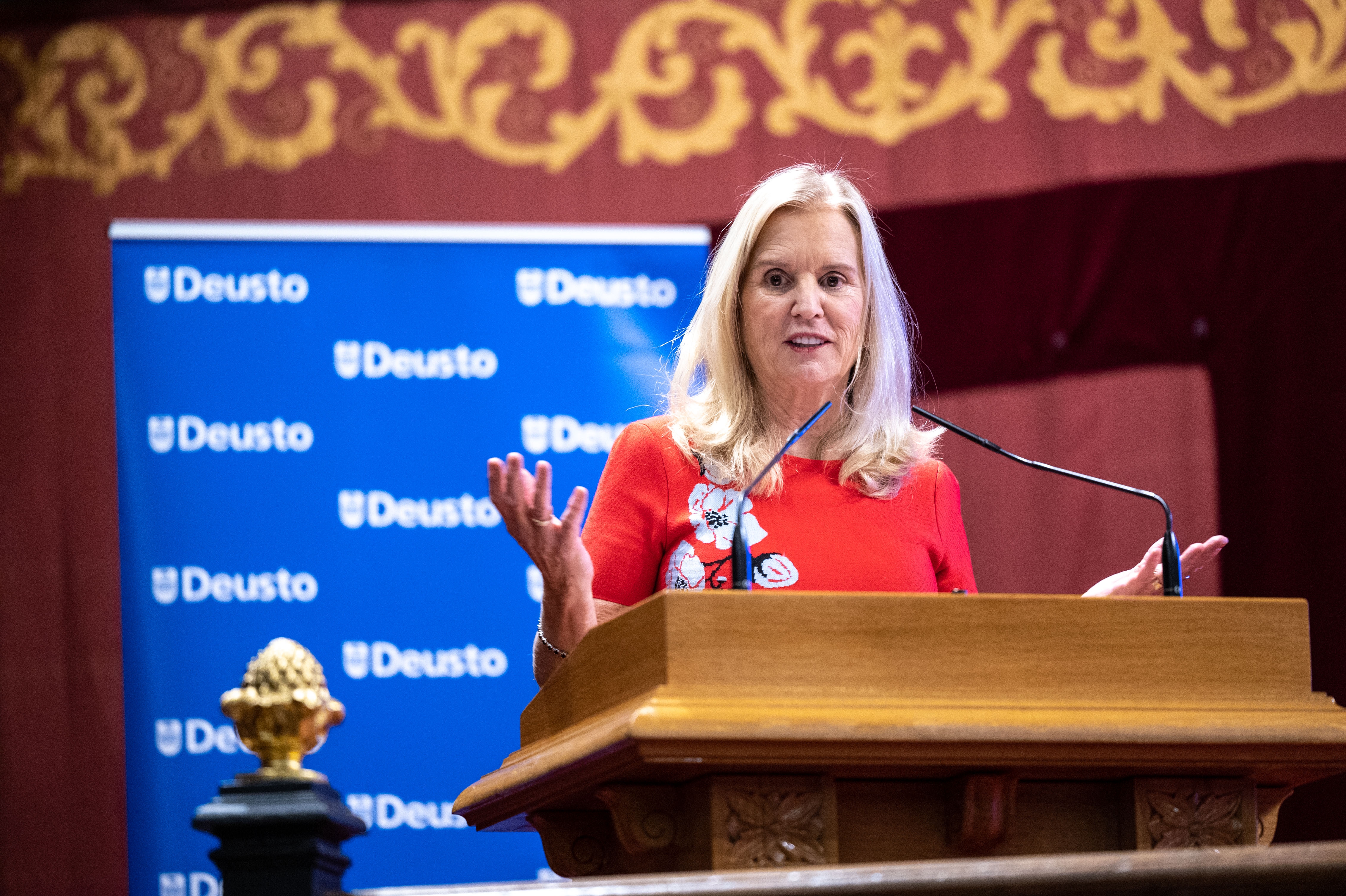 Person speaking at a podium with a "Deusto" banner in the background. They are gesturing with their hands, wearing a professional outfit