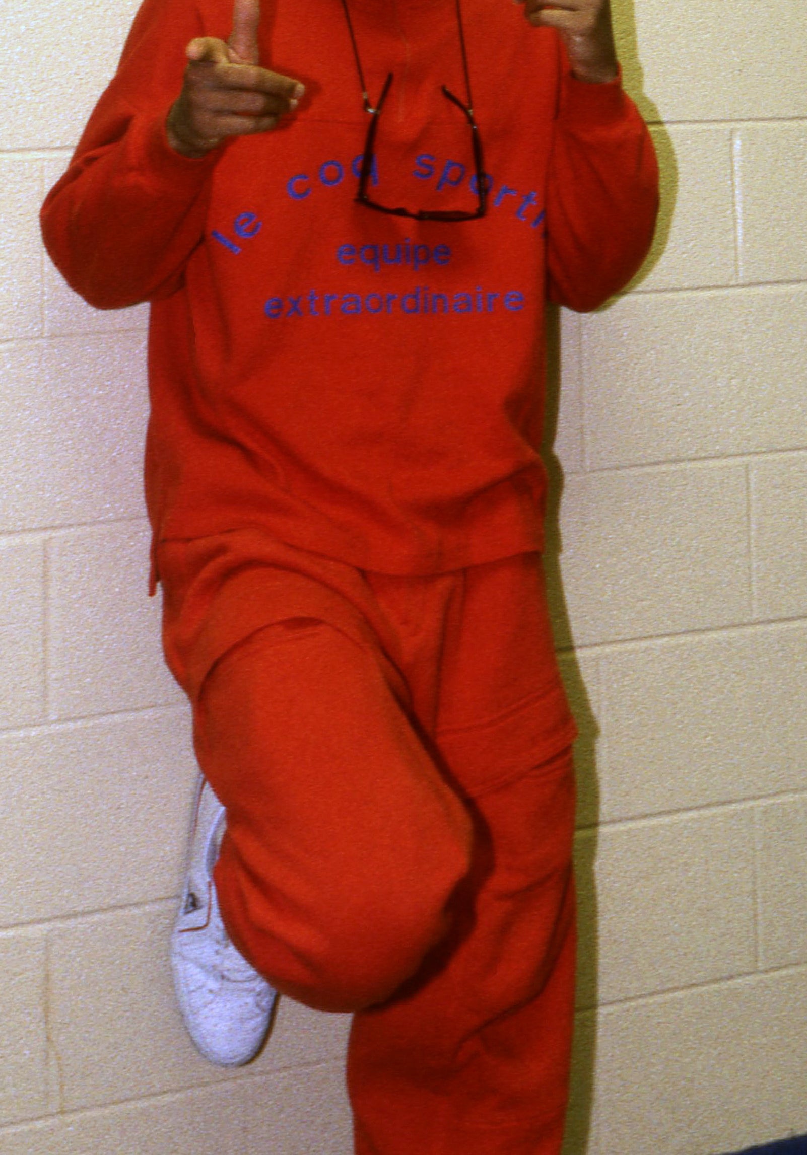 Celebrity in a casual red athletic outfit poses playfully, leaning against a wall with a finger-gun gesture, wearing white sneakers