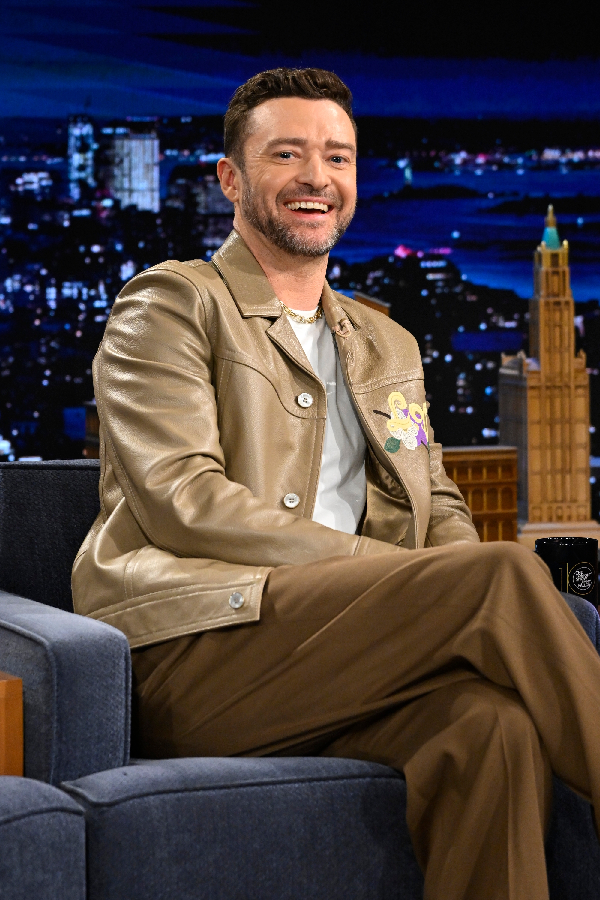 Person seated on a talk show set wearing a stylish tan leather jacket and pants, smiling
