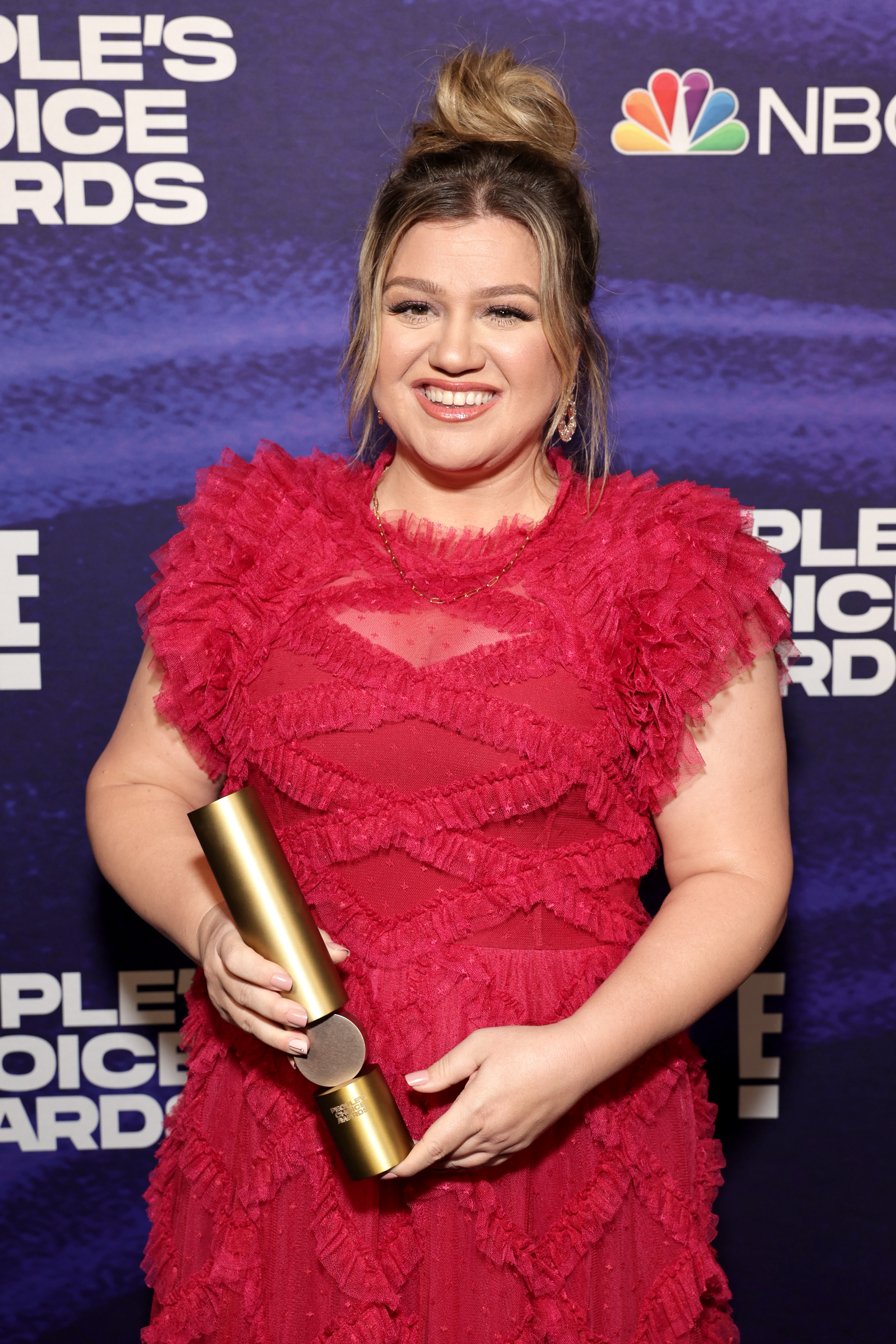 Person in a ruffled dress smiling and holding a gold award on a red carpet, with a People's Choice Awards backdrop