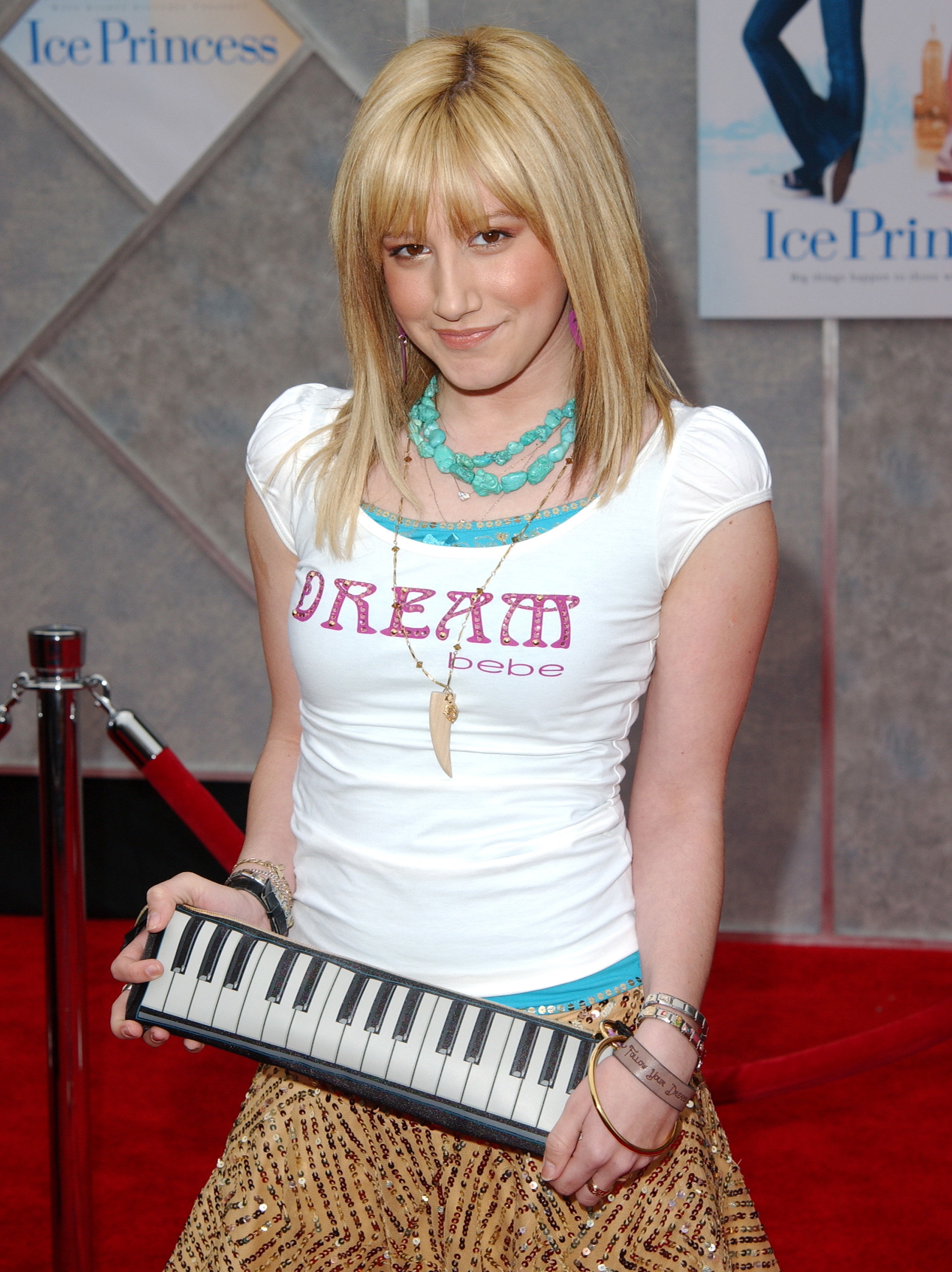 Person on red carpet holds mini keyboard, wearing graphic tee and layered necklaces, smiling at camera