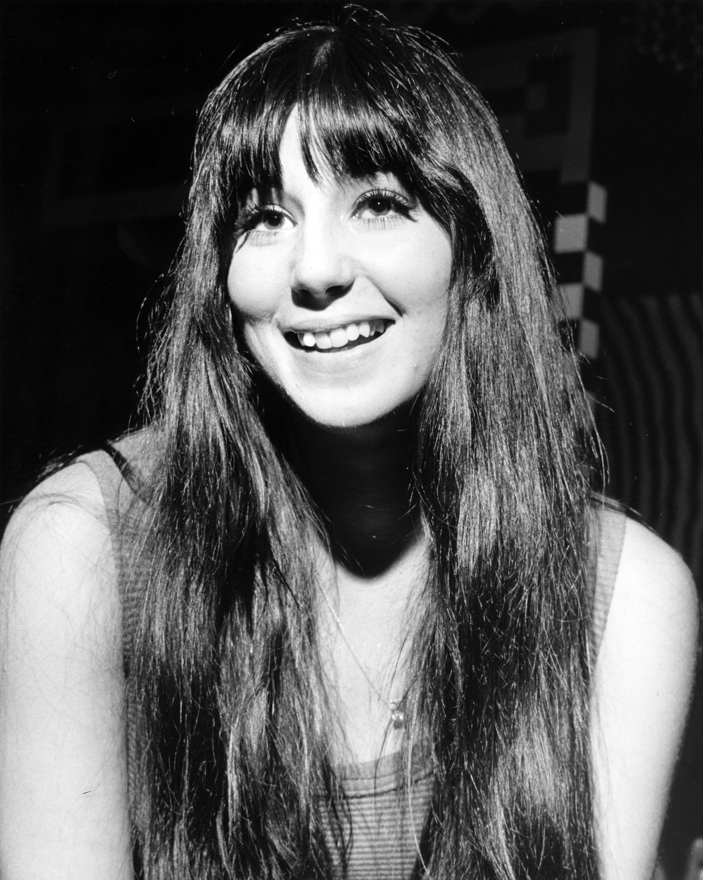 A woman with long hair and bangs smiles warmly at the camera in a casual setting