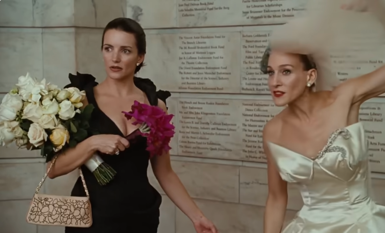 Two people at a wedding, one in a white strapless gown with a veil, surprised expression, the other in a black dress holding flower bouquets