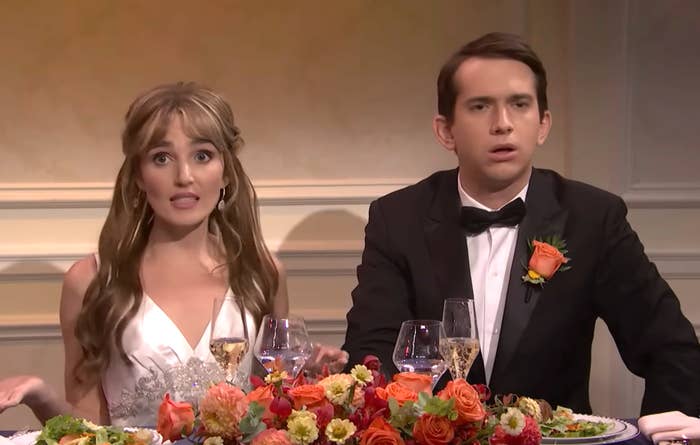 A couple at a wedding reception sits at a table adorned with colorful floral arrangements. The woman wears a white dress; the man is in a tuxedo