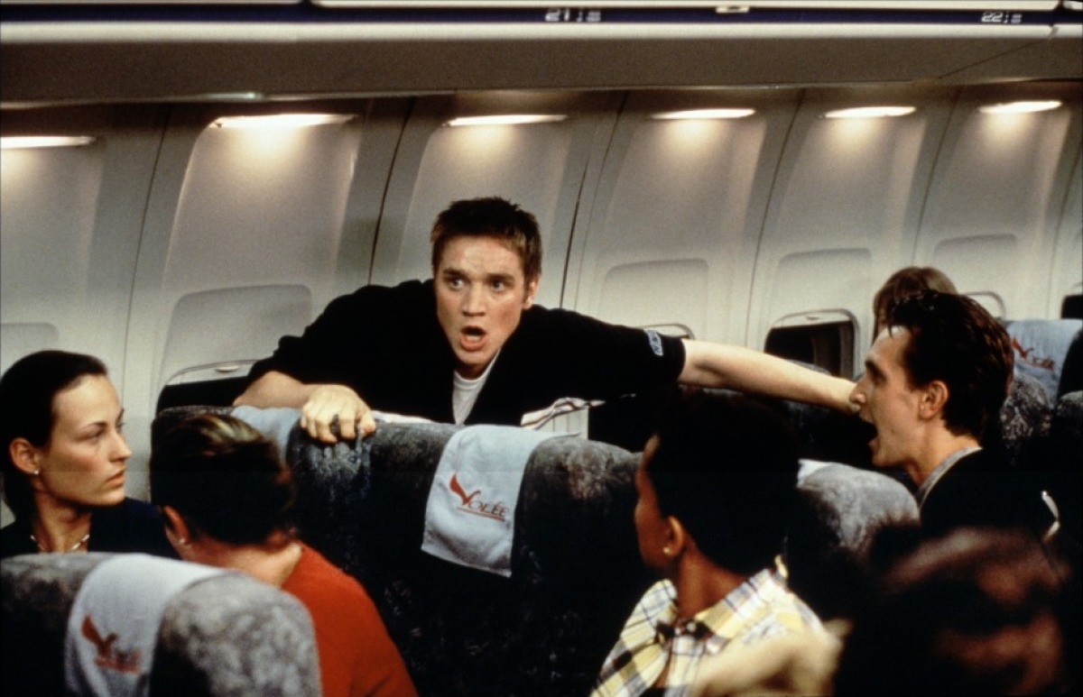 Person stands in airplane aisle, appearing surprised and animated, surrounded by seated passengers in a movie scene