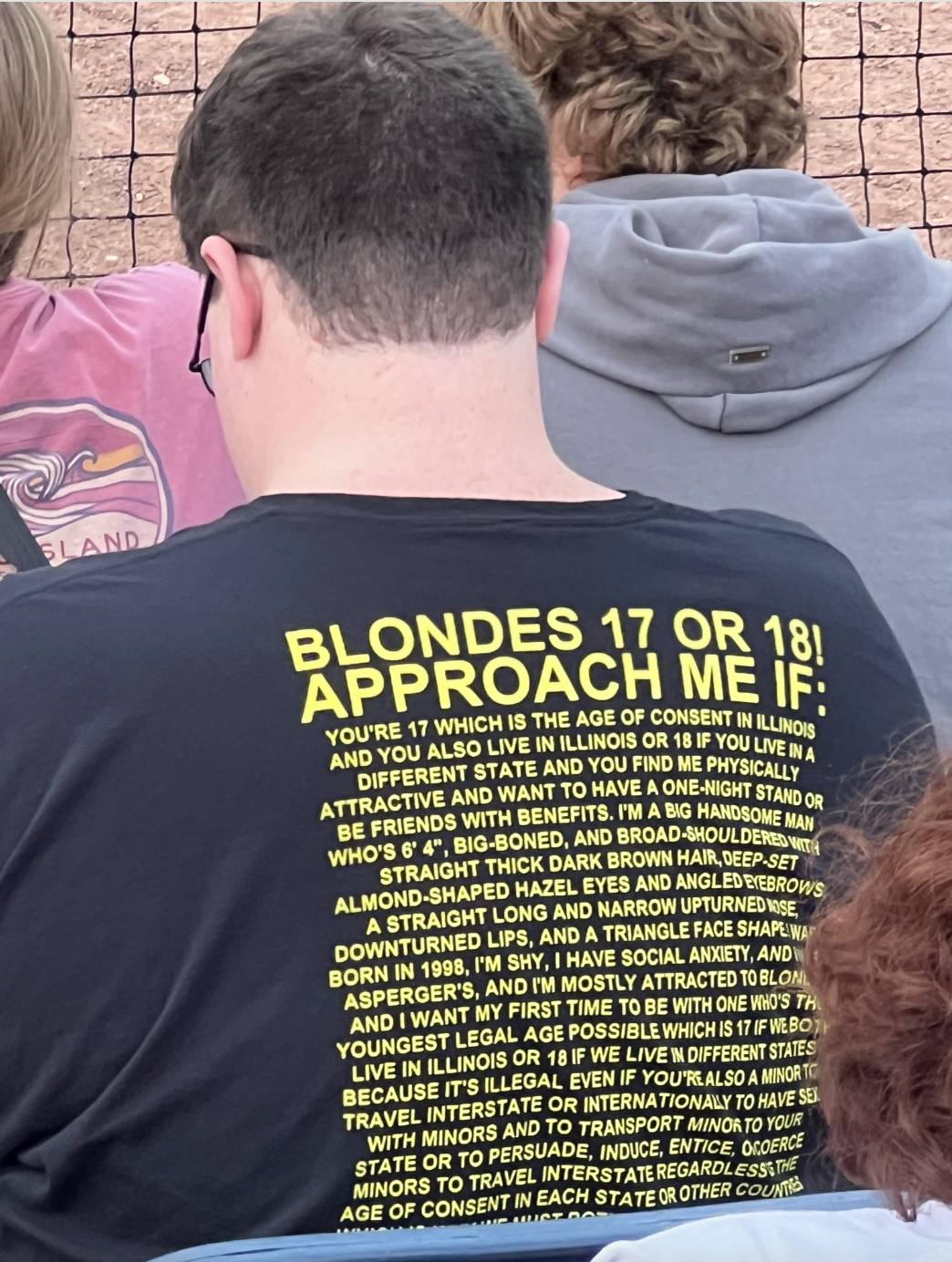 Person wearing a shirt with large text about dating preferences, seen from behind, in a casual outdoor setting with other people