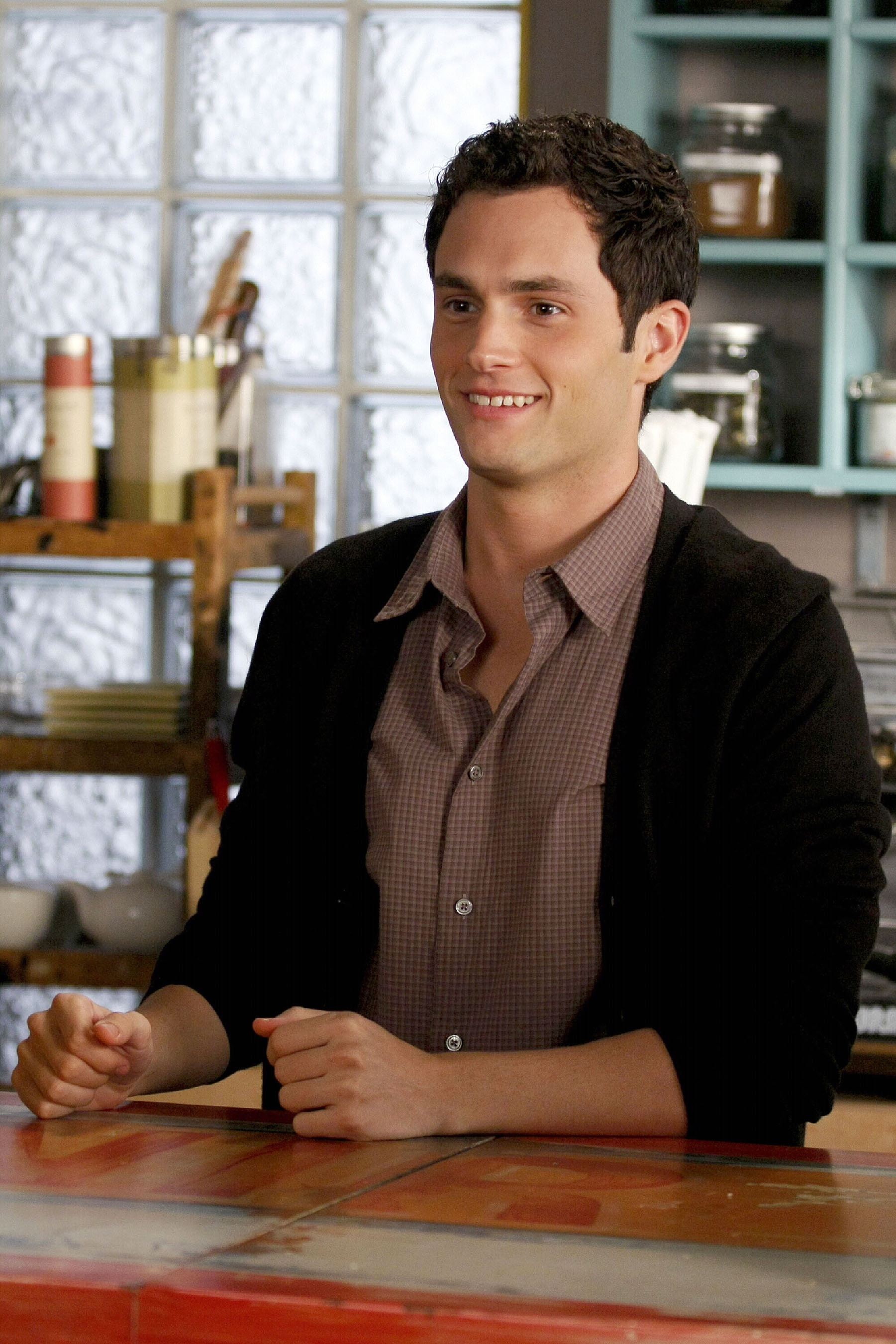 Person in casual attire with a checkered shirt and dark cardigan, smiling while seated at a kitchen counter