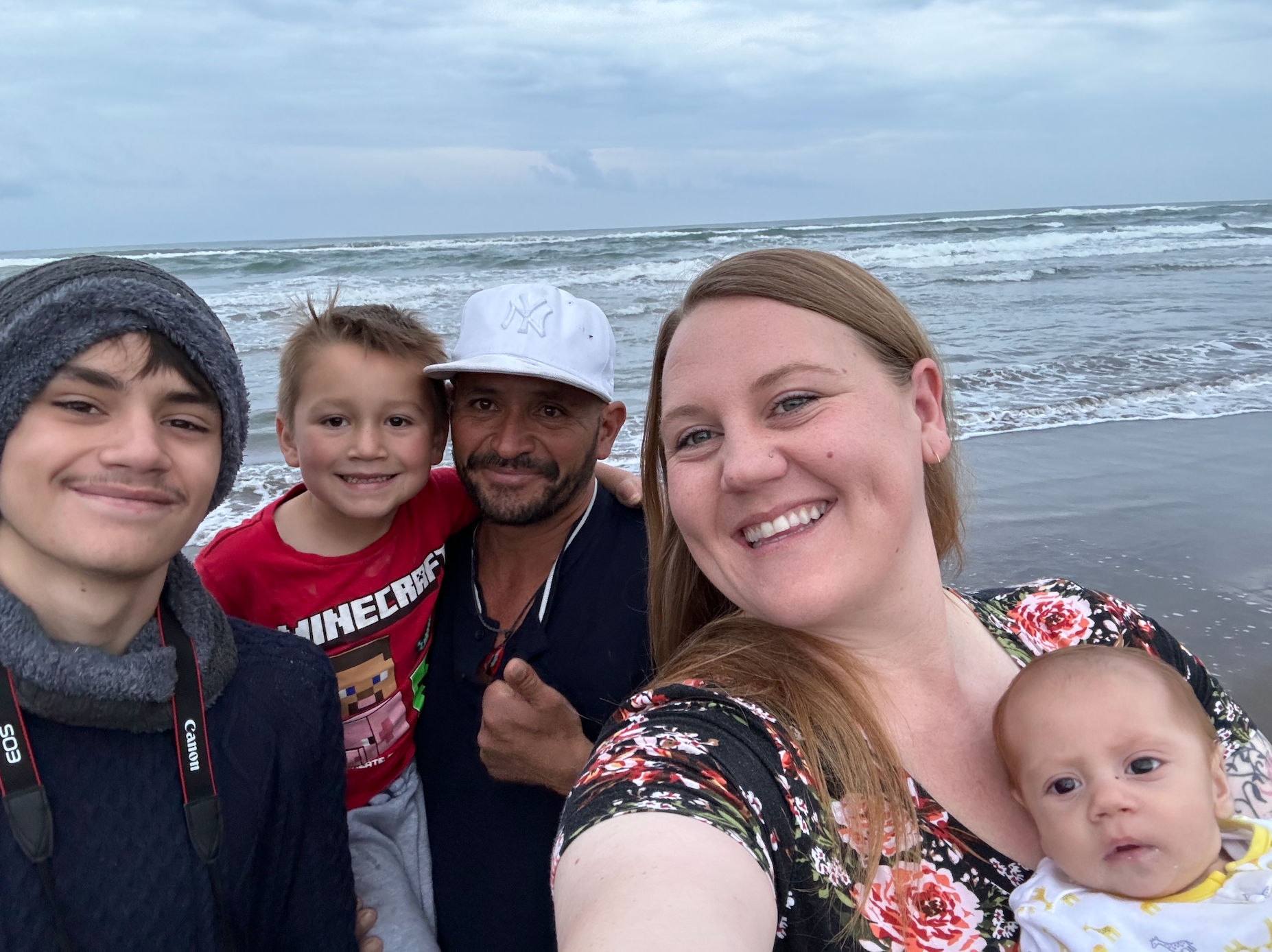Family of five smiling for a selfie on a beach, with waves in the background