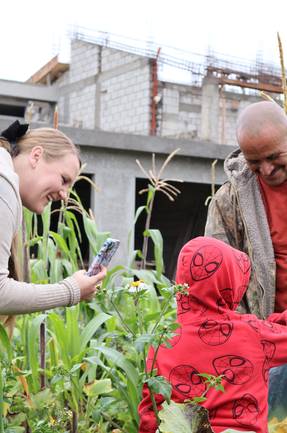 A woman takes a photo of a child in a Spider-Man hoodie, with a man nearby in a rural garden setting