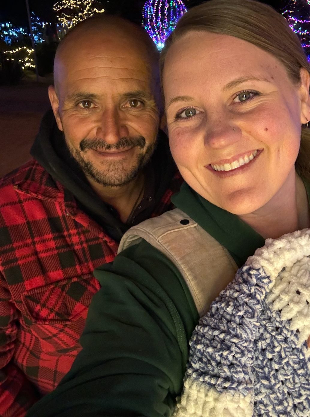 Man in plaid jacket and woman holding a blue and white knit blanket smile outdoors at night, surrounded by festive lights