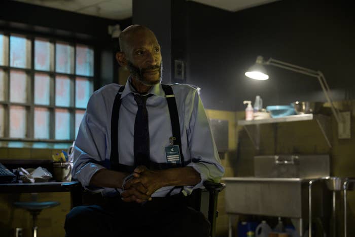 A man sits in a dimly lit room, wearing a dress shirt with suspenders and an ID badge, suggesting a scene from a TV show or movie