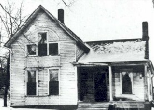 Old, two-story wooden house with a porch, surrounded by bare trees, suggesting a winter setting