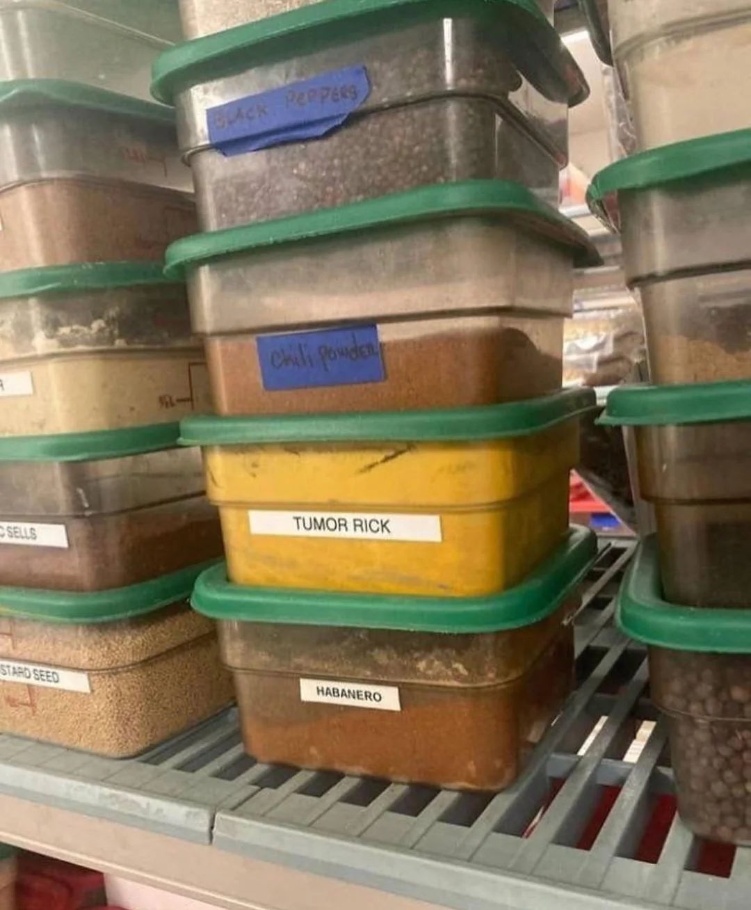 Stacked containers labeled with various spices, including "Chilli Powder," "TUMOR RICK," and "HABANERO," on a shelf in a storage area