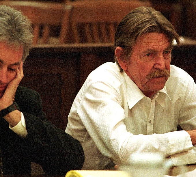 Two men sit at a table in a courtroom, appearing serious. One is in a white shirt, the other in a suit with a patterned tie