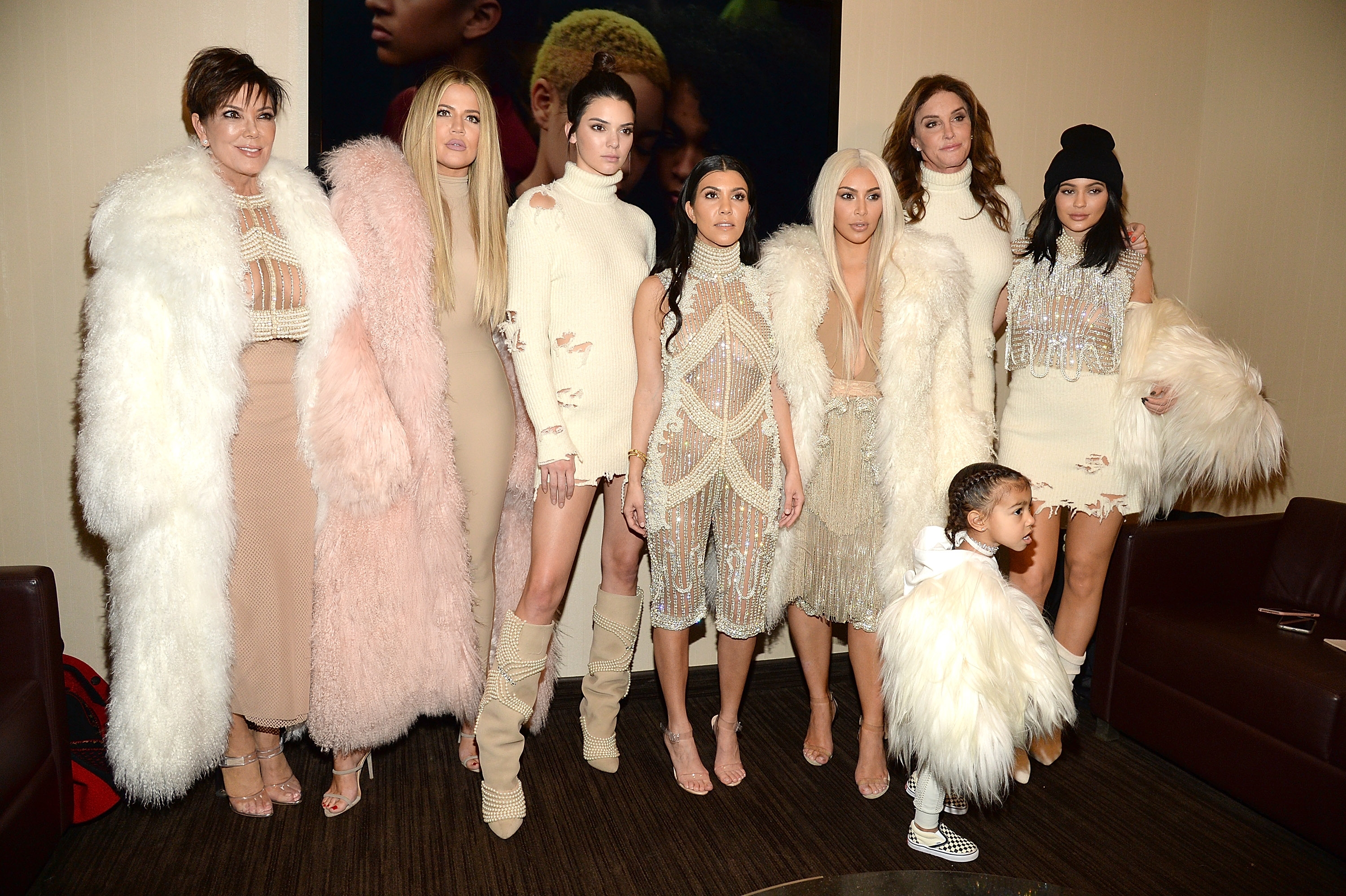 Kardashians poses in a room wearing stylish, coordinated outfits with fur and sparkly details. A child stands in front