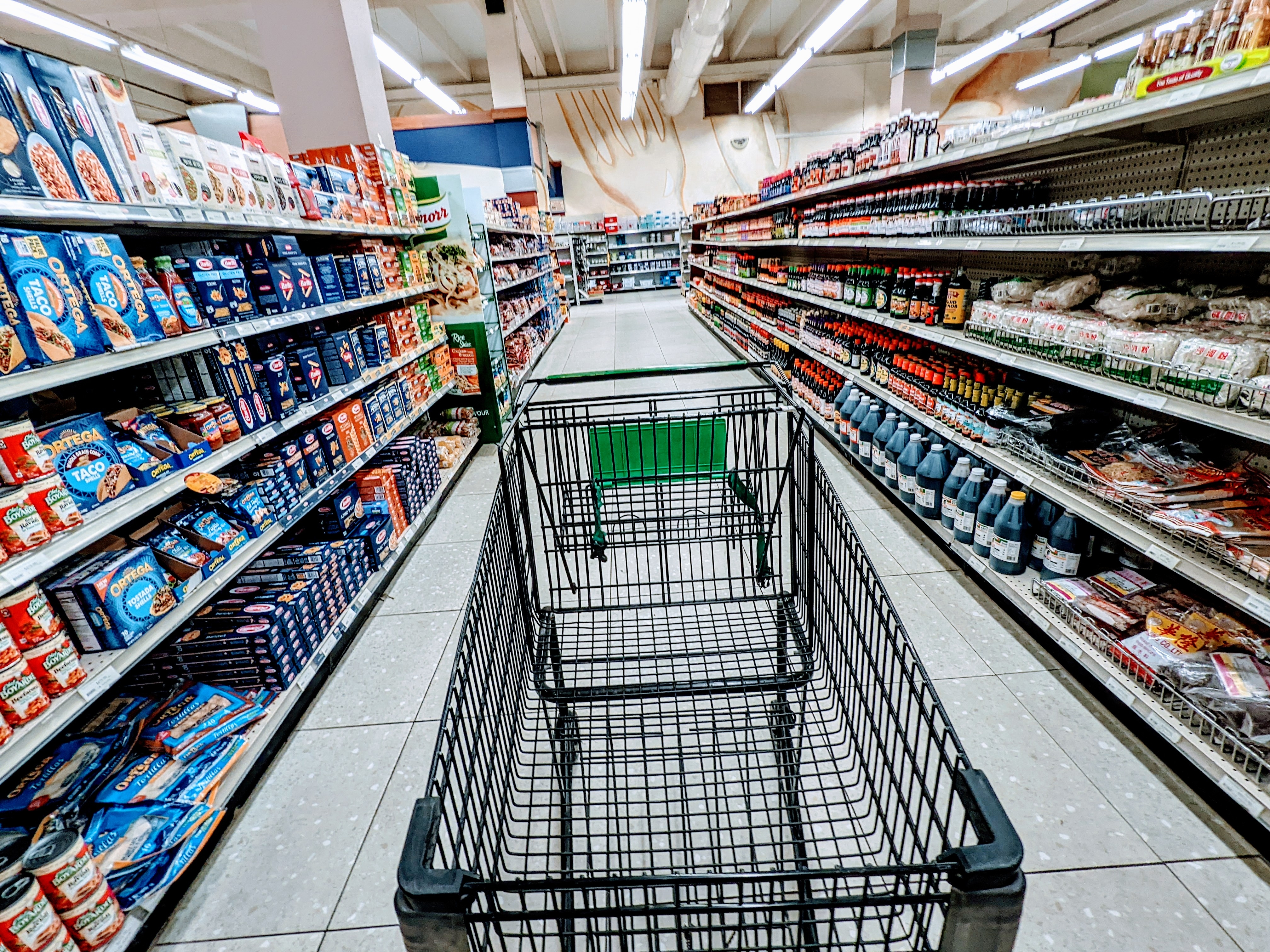 Shopping cart in an empty supermarket aisle, surrounded by stocked shelves with various packaged food and beverage items
