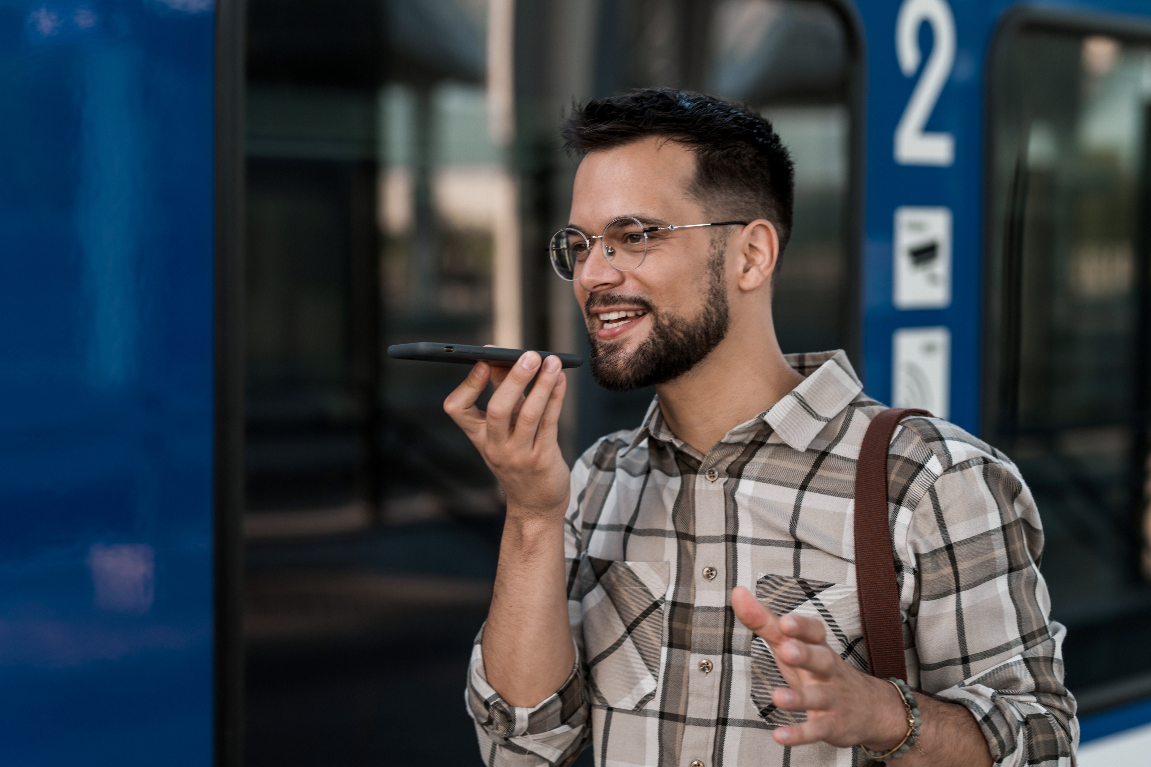 Person in a plaid shirt speaks into a smartphone outside, standing near a modern blue tram
