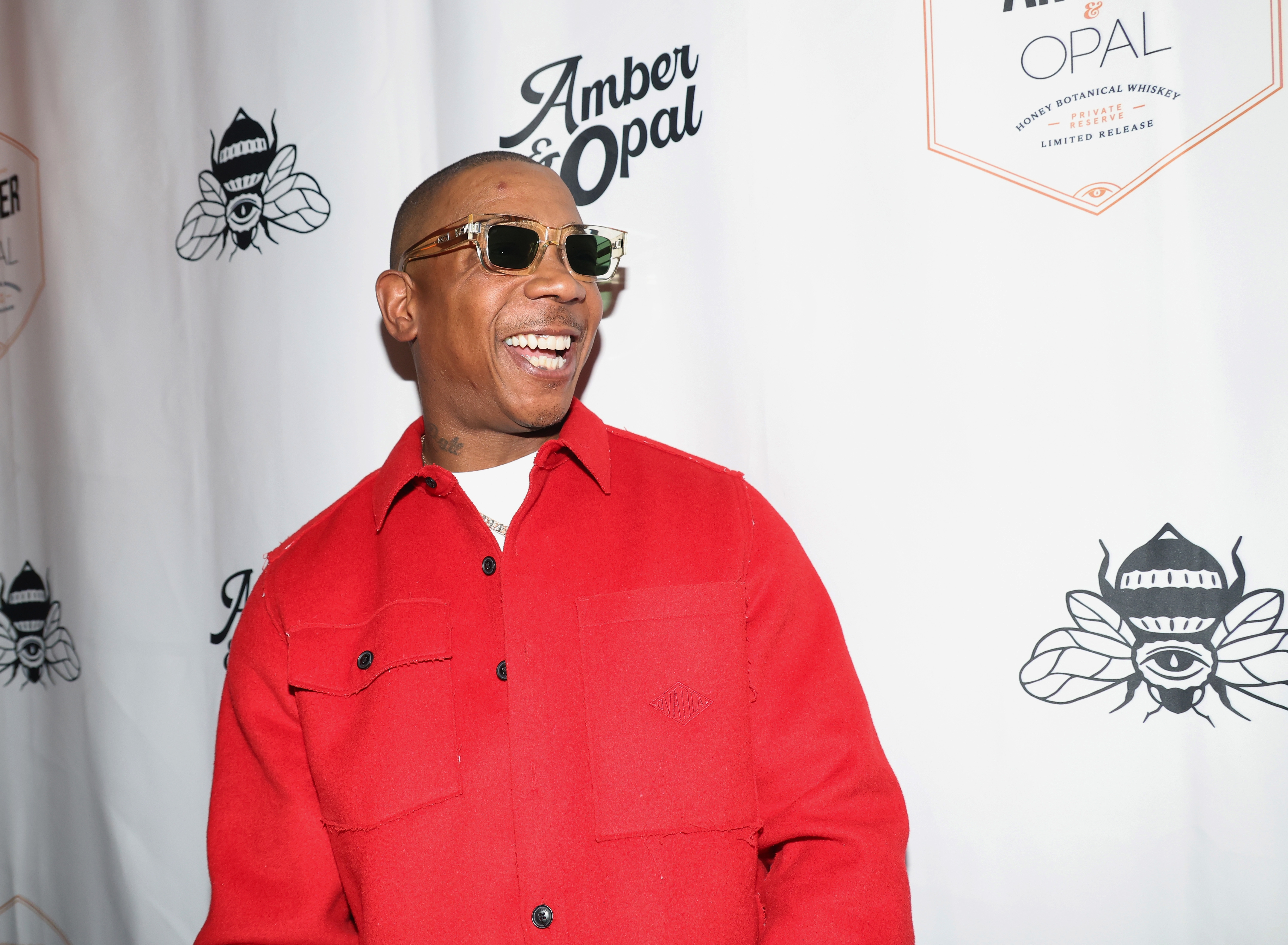 Ja Rule in sunglasses and red shirt smiles at a wall with "Amber & Opal" logos