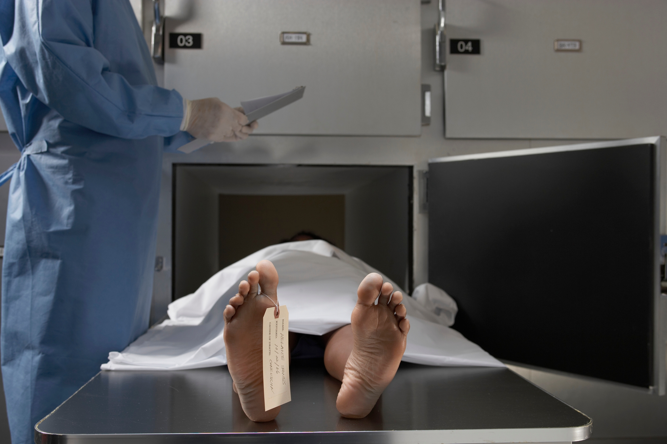 A covered body on a morgue table with a tag on the toe; a medical professional stands nearby holding a clipboard
