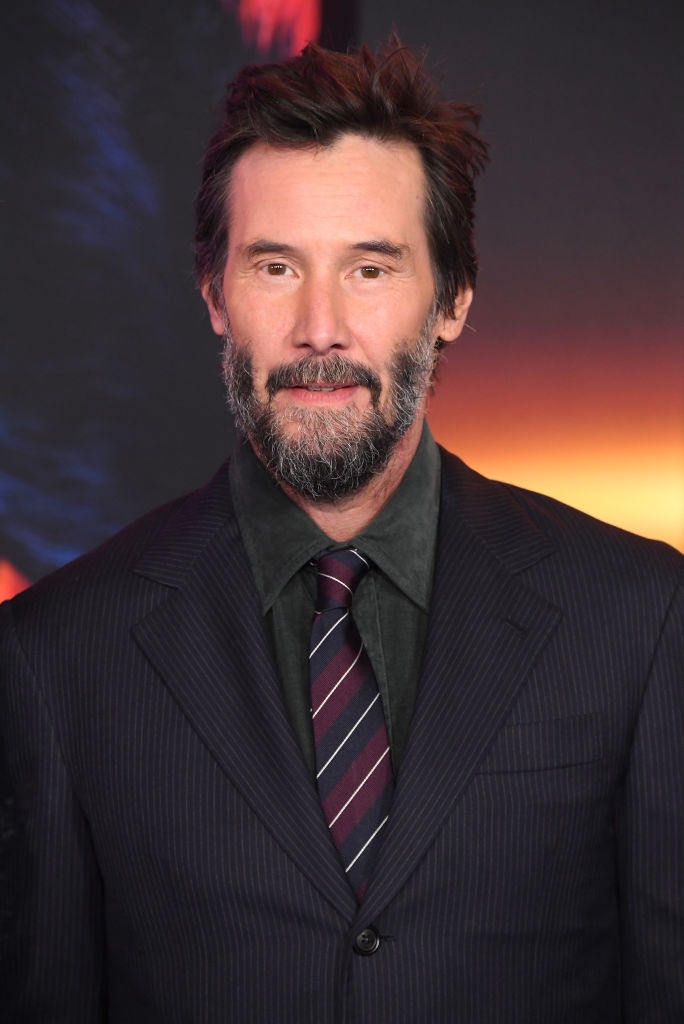 A man in a pinstripe suit and tie with a beard stands against a blurred background on a red carpet