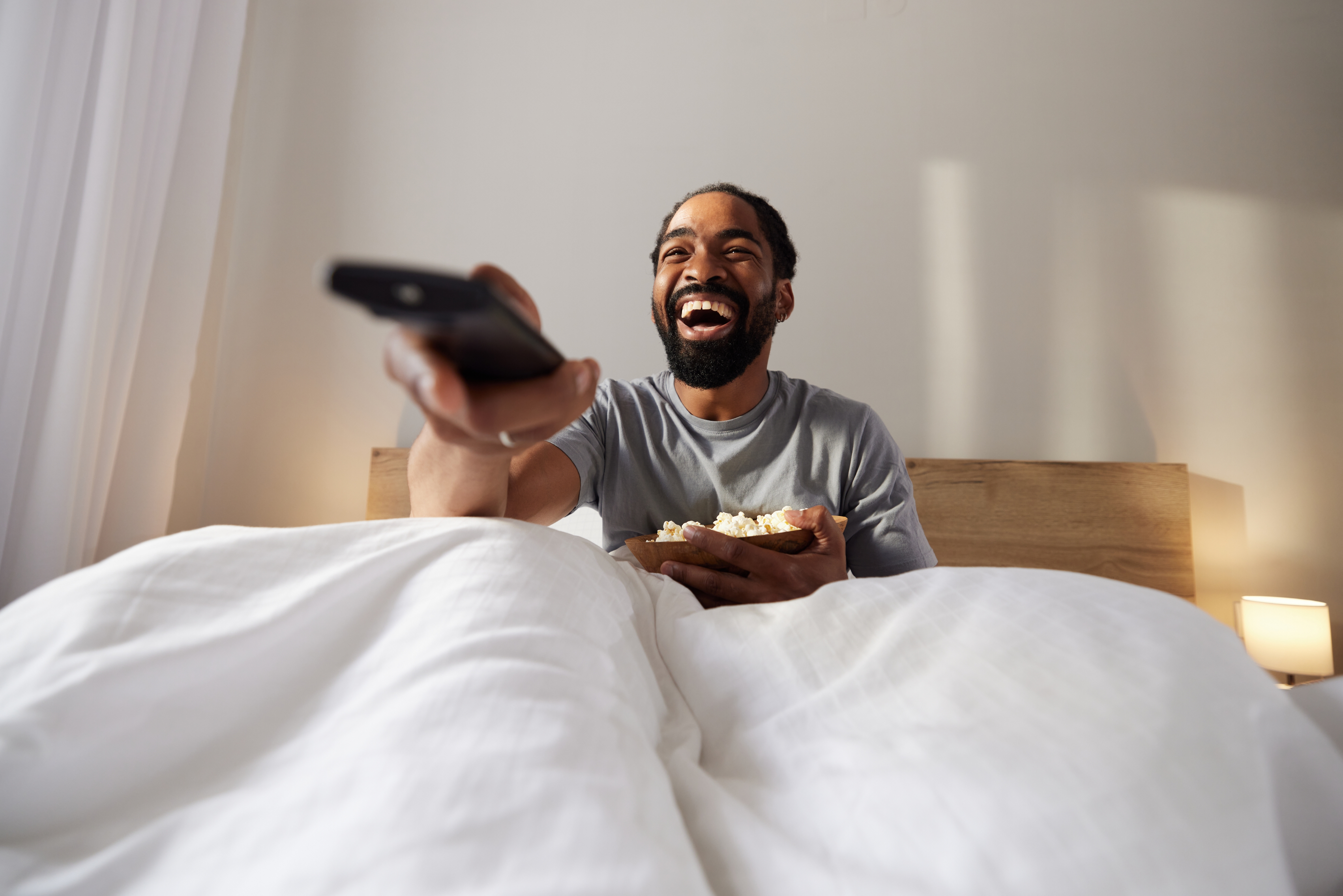 A man in bed with a remote, laughing while holding a bowl of popcorn, suggesting a relaxed and enjoyable moment