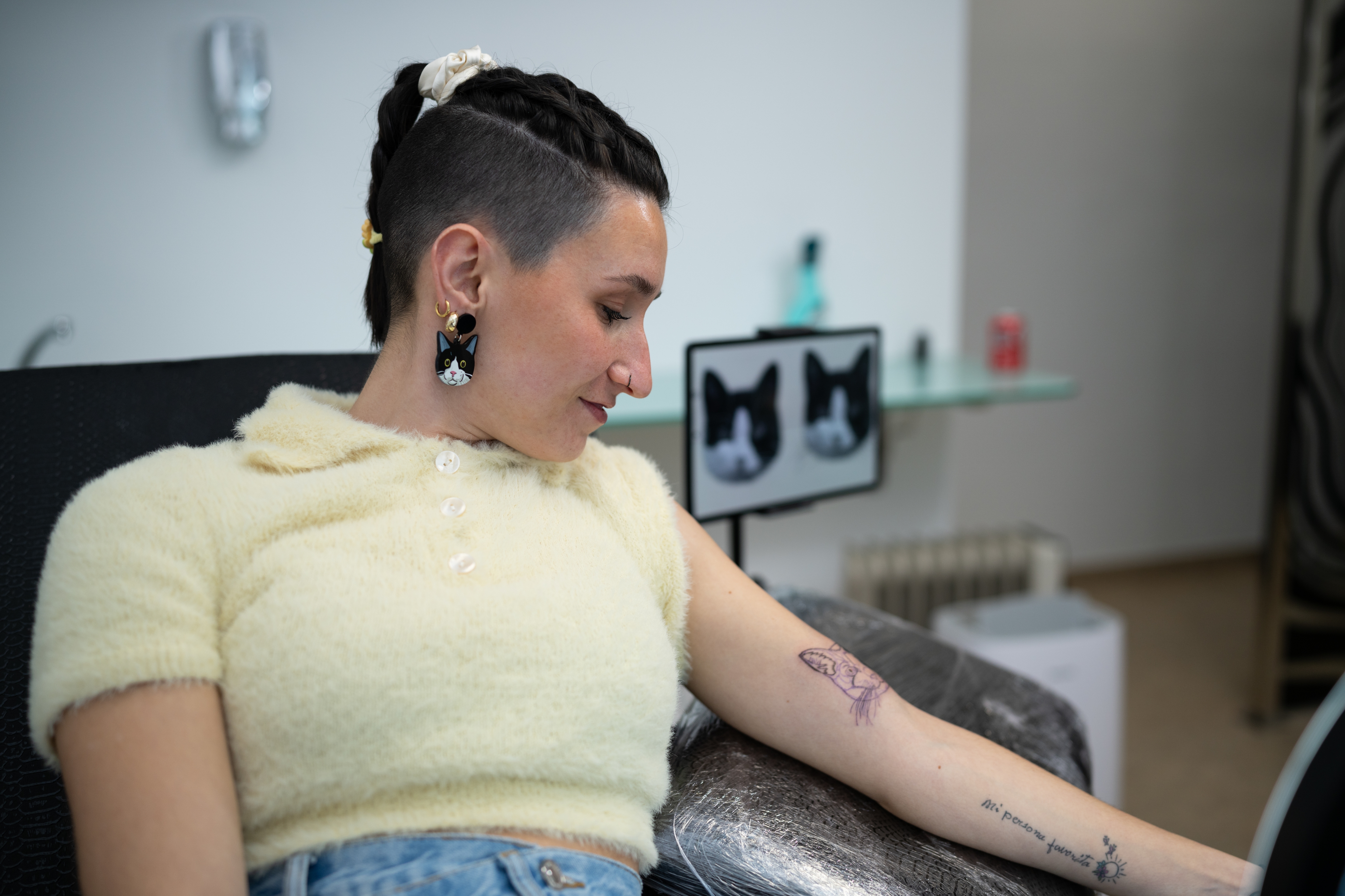 Person sitting, admiring a fresh forearm tattoo showing a butterfly design. They wear a short-sleeve top and cats are visible in the background decor