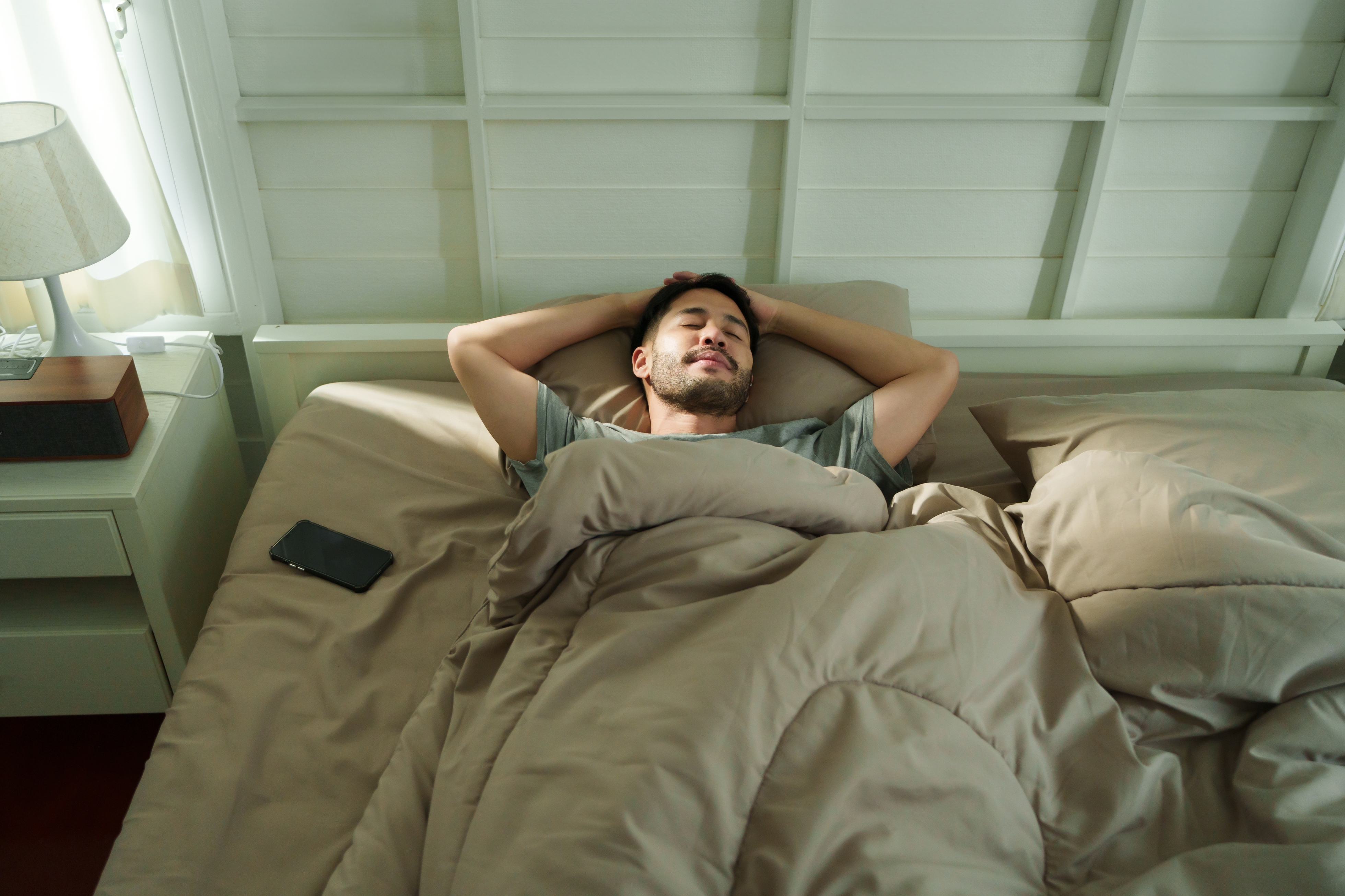 Man lying in bed with arms behind head, eyes closed, appearing relaxed. Smartphone on bedside table. Cozy bedroom setting