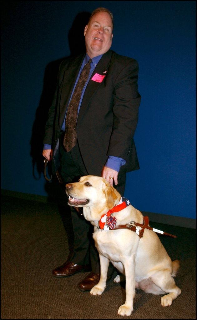 Man in a suit smiling, standing with a guide dog wearing a harness and a patriotic scarf
