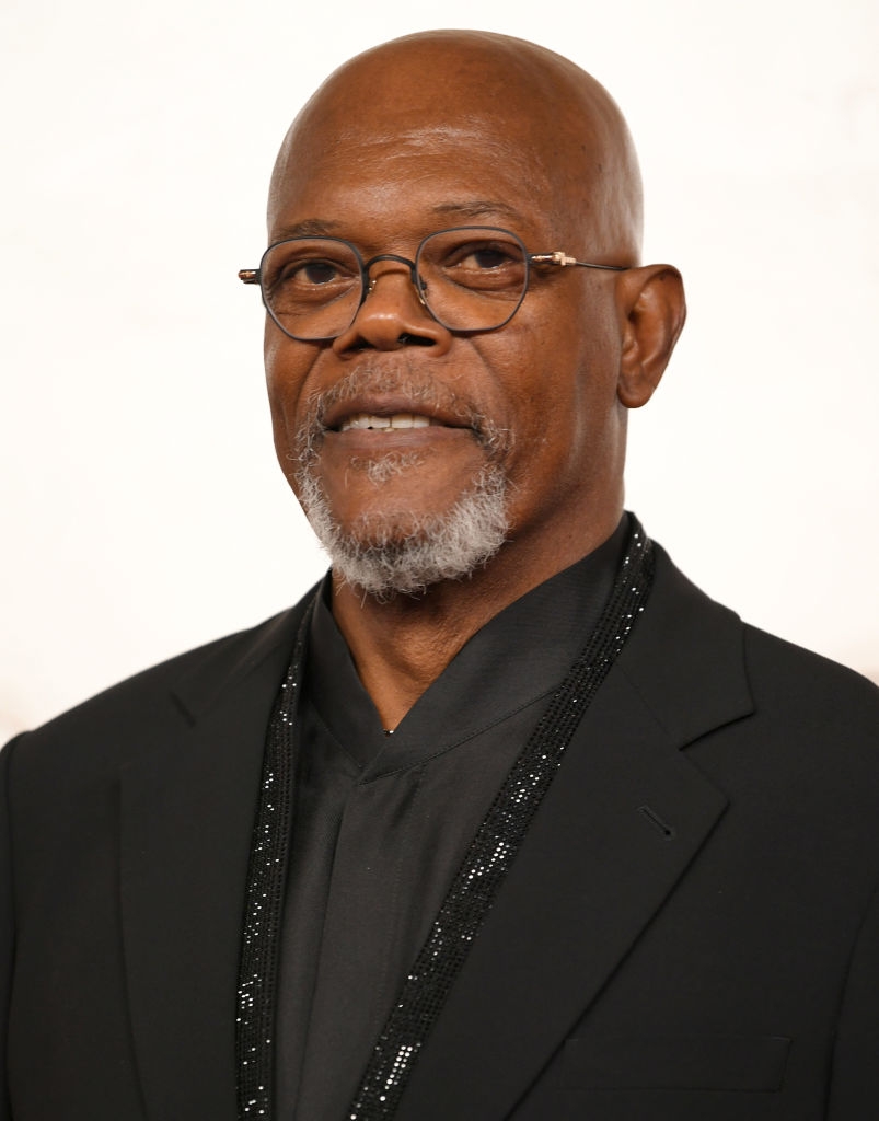 Man in a formal black suit and glasses poses against a neutral background